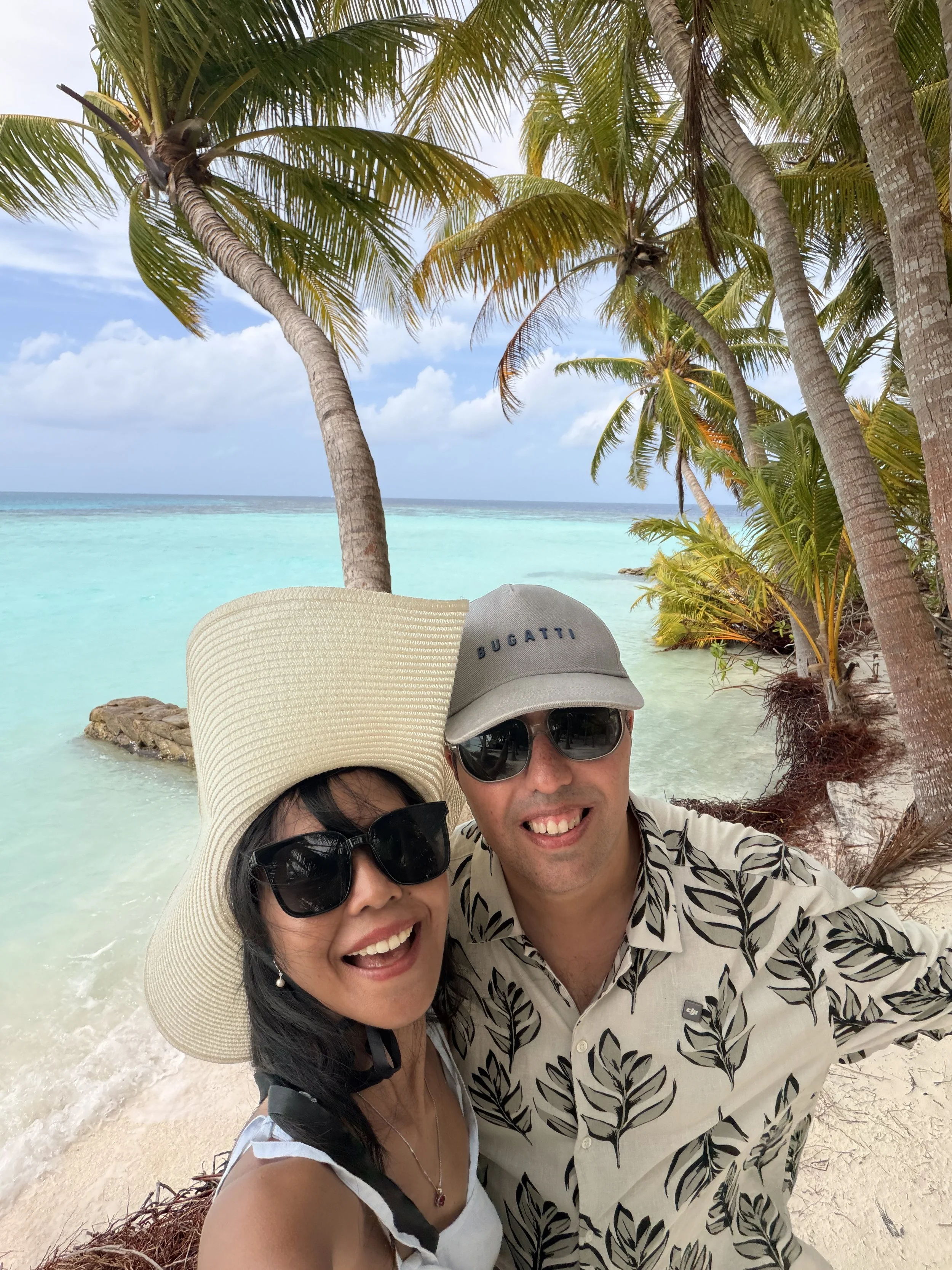 A smiling couple taking a selfie on a tropical beach with turquoise water, palm trees, and partly cloudy sky in the background.