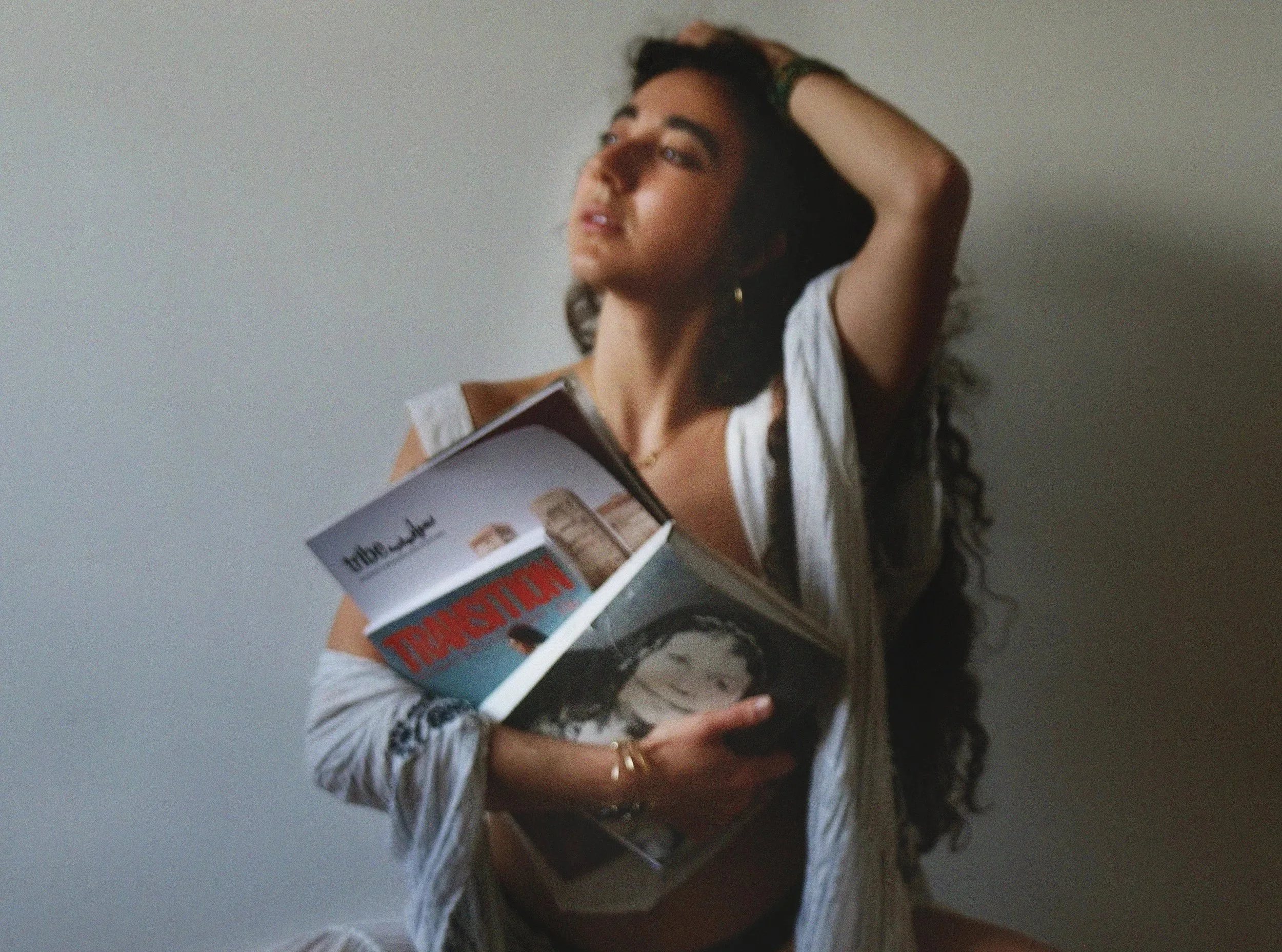 A woman holding a book titled 'The Subtle Art of Not Giving a F*ck' while standing against a plain wall. She has long curly hair, is wearing a white top, and has her left arm raised with her hand resting on her head.