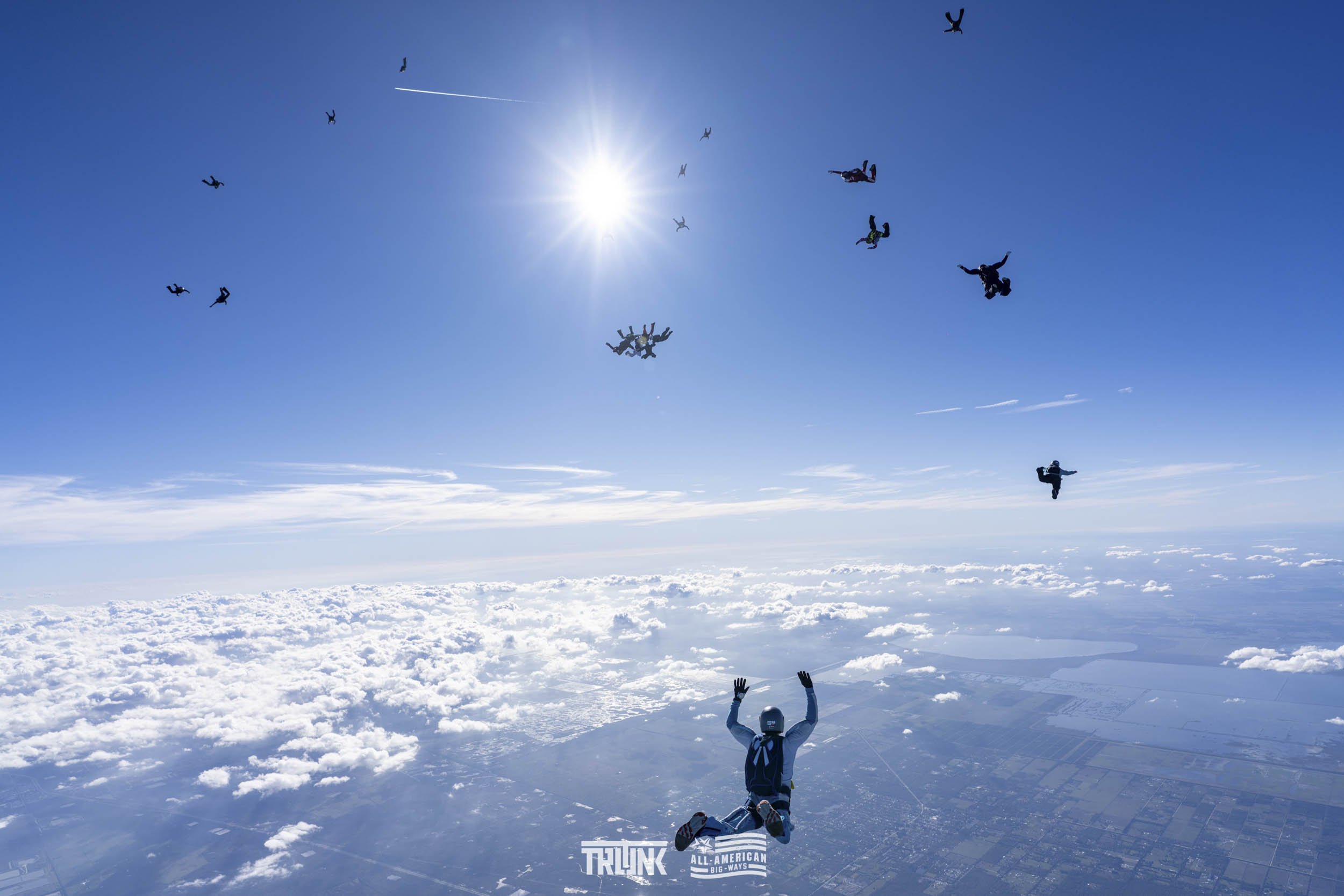 Skydive group freefalling over clouds with clear blue sky and bright sun, one person with arms raised in the foreground and others spread out in various positions.