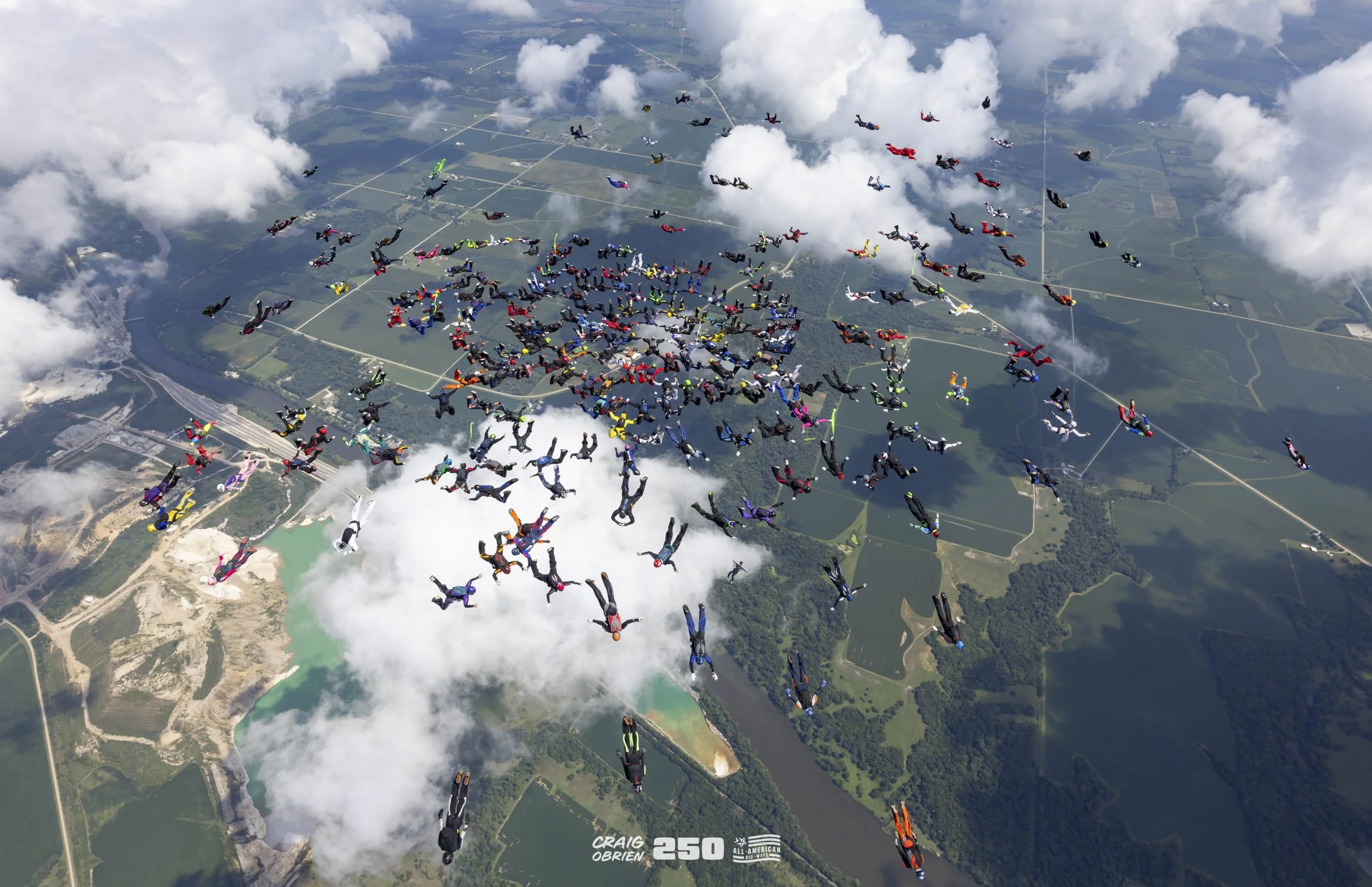 Aerial view of hundreds of skydivers in colorful parachutes mid-air over a landscape with lakes and farmland, with some clouds in the sky.