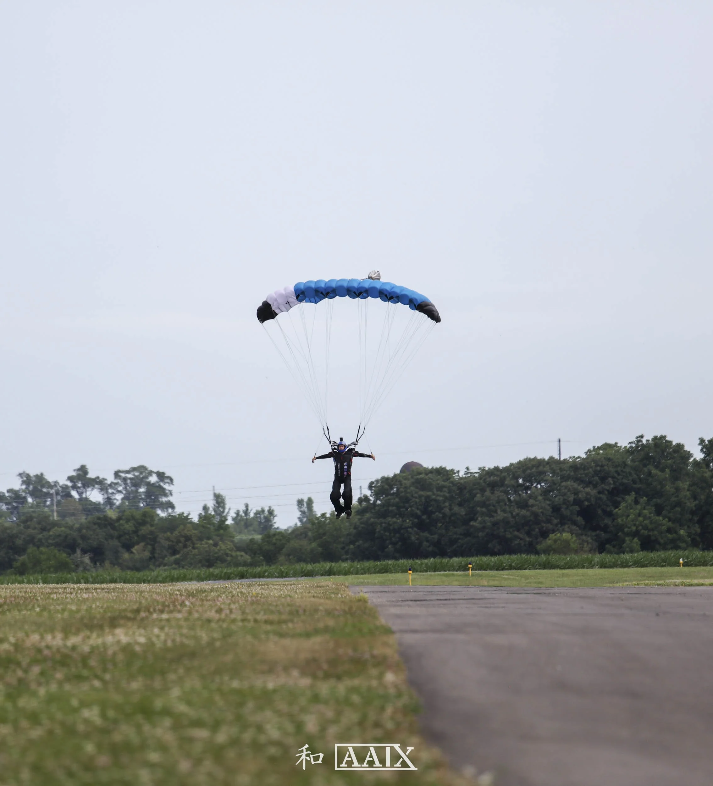 Person landing with parachute on grassy field adjacent to runway, surrounded by trees, overcast sky.