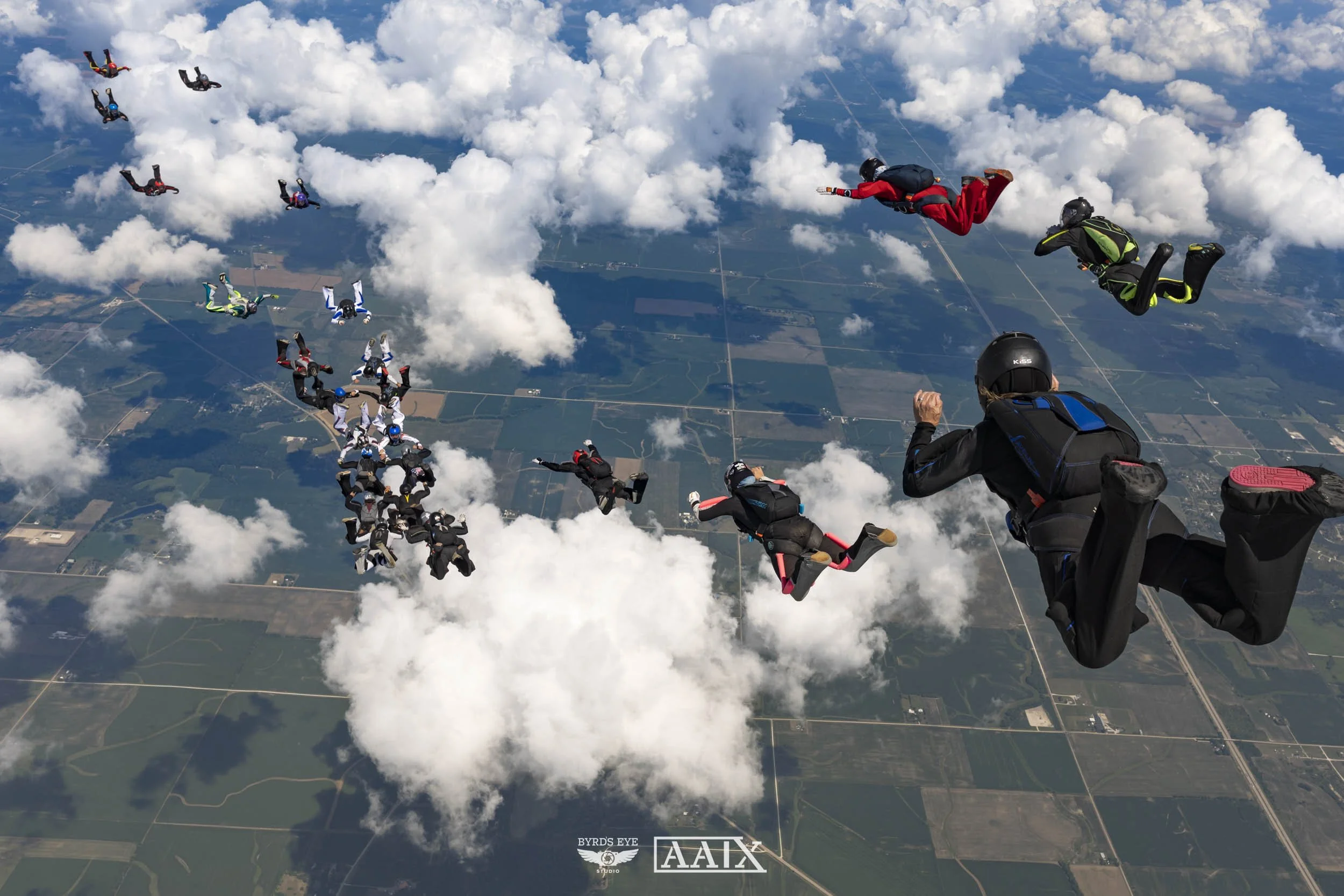 Skydivers in free fall above the clouds, with a landscape of fields and roads below.