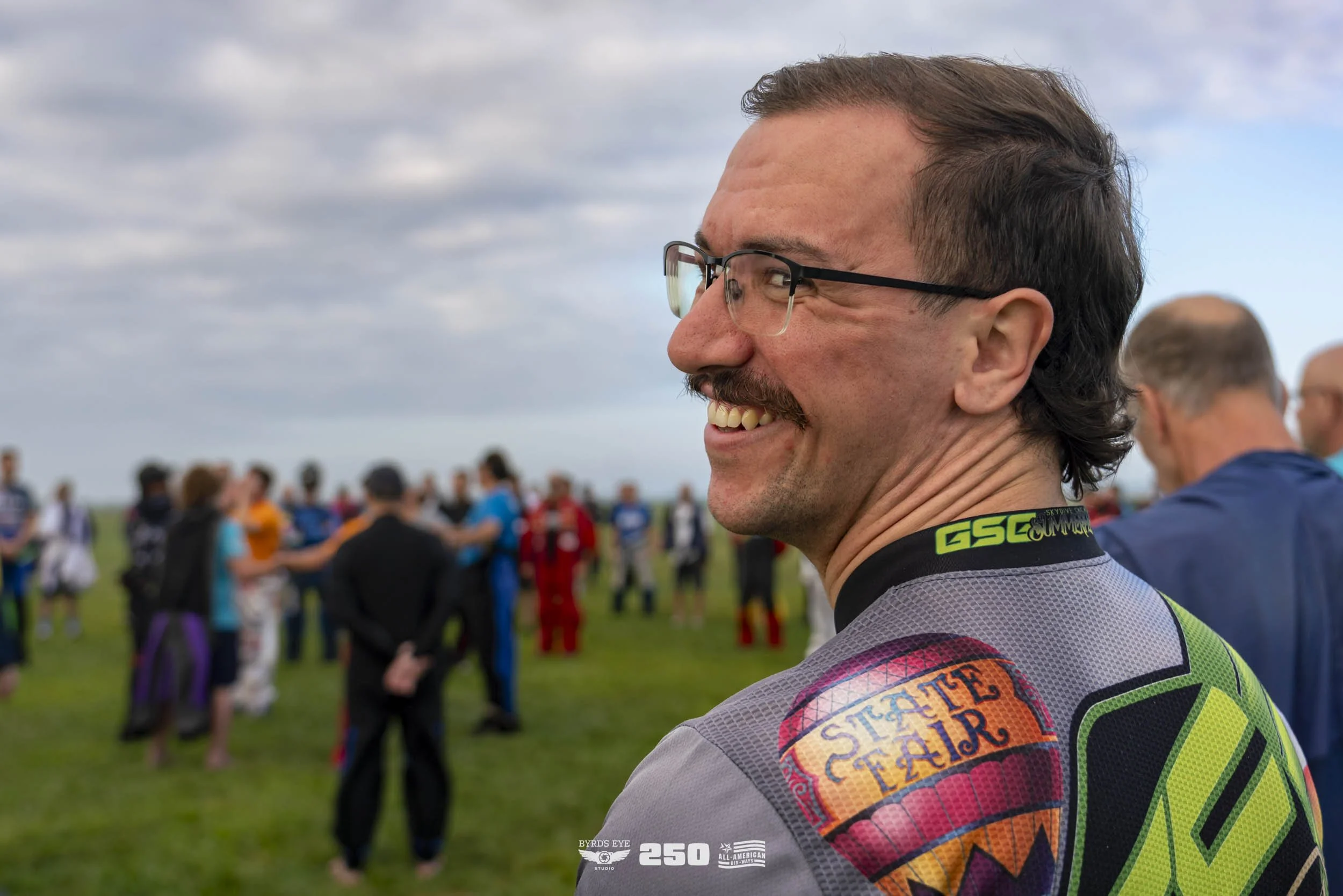 A man smiling and wearing glasses and a cycling jersey with a 'State Fair' logo, amid a group of people outdoors on a grassy field.