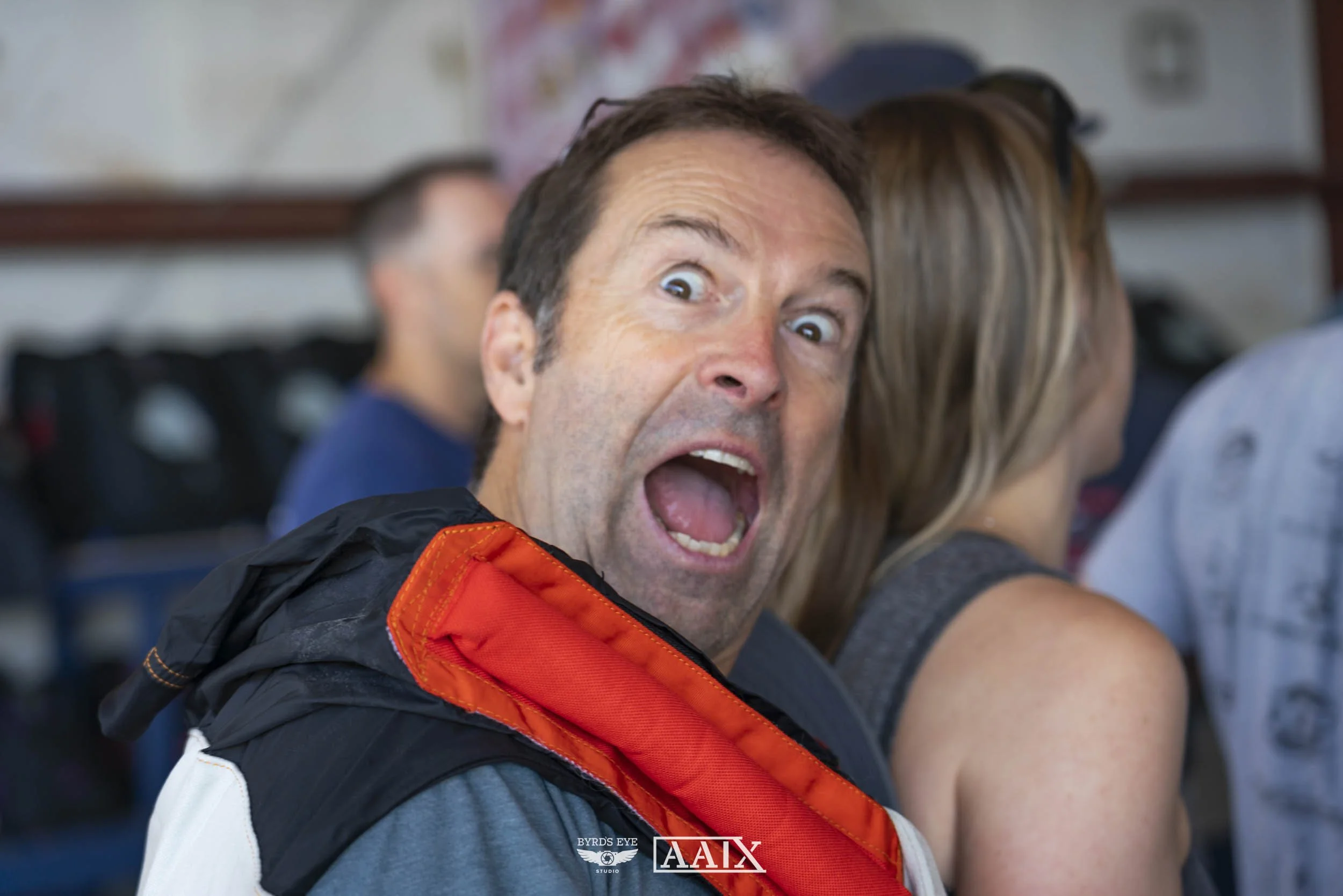 A man with short dark hair, wearing a grey shirt and a red and black jacket, making a surprised or shocked expression with his mouth open wide and eyes wide open.