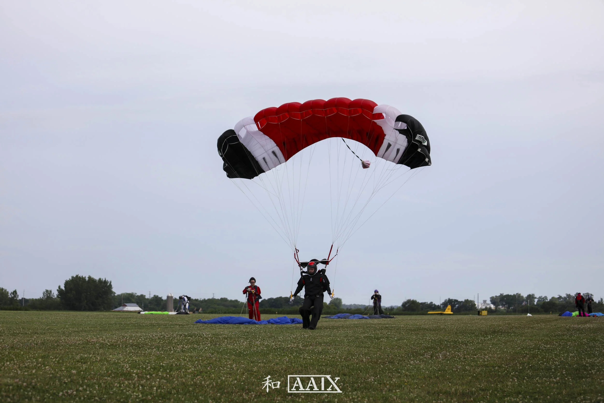 Person in a helmet and suit taking off for a parachute jump on a grassy field, with others preparing parachutes in the background.