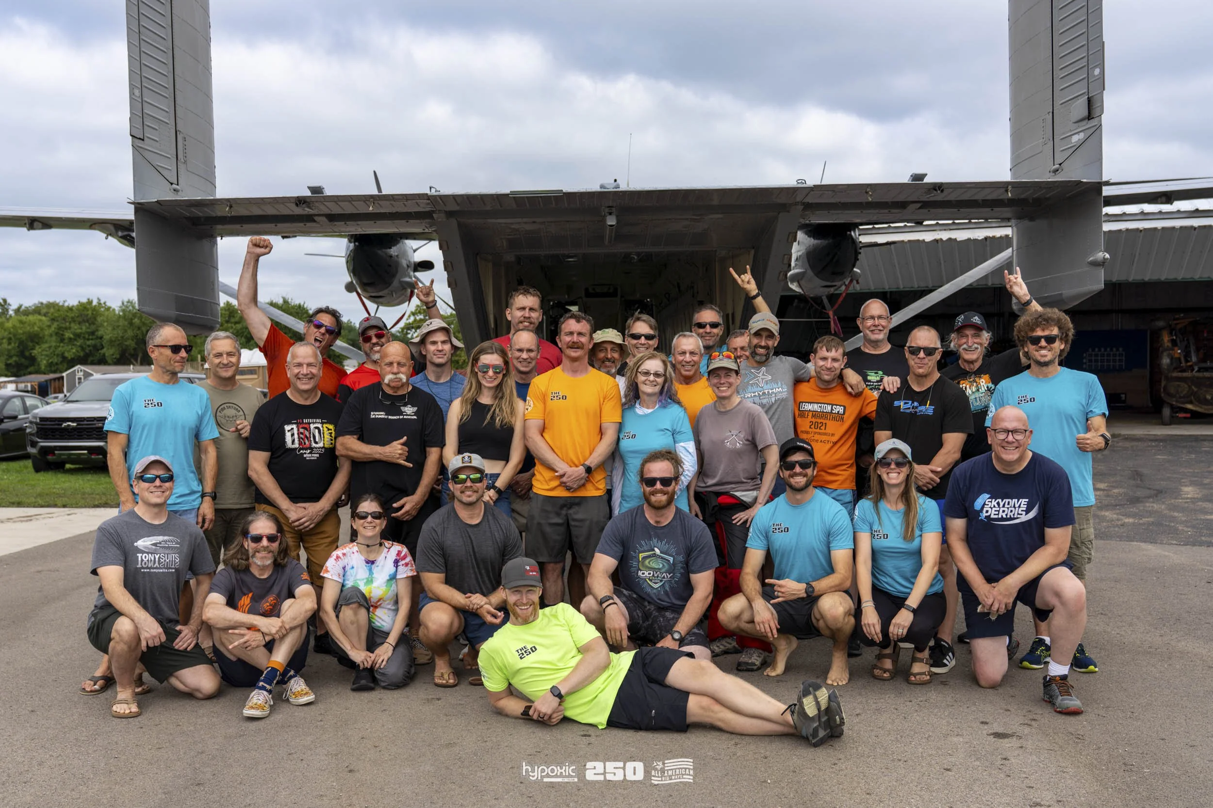 Group of people standing and sitting in front of a military transport aircraft, some raising their hands or making gestures, on a paved surface during daytime.
