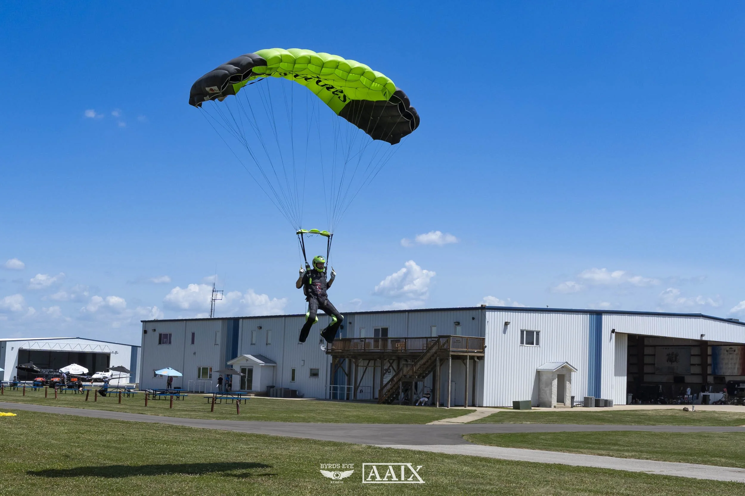A person in skydiving gear descending with a green and black parachute in clear blue sky near an industrial building.