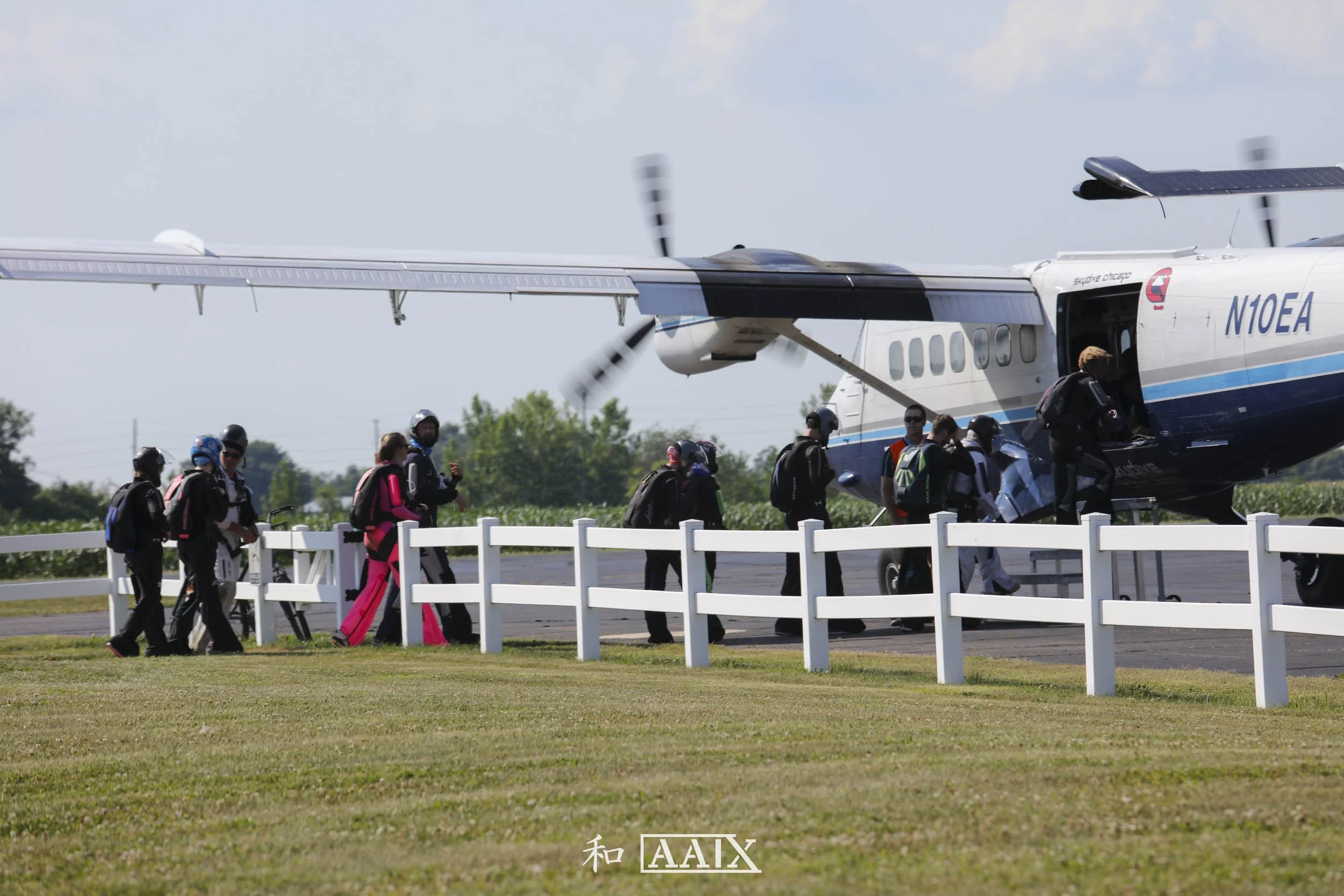 A group of people walking towards a small aircraft on an airport tarmac, with a clear blue sky and green trees in the background.