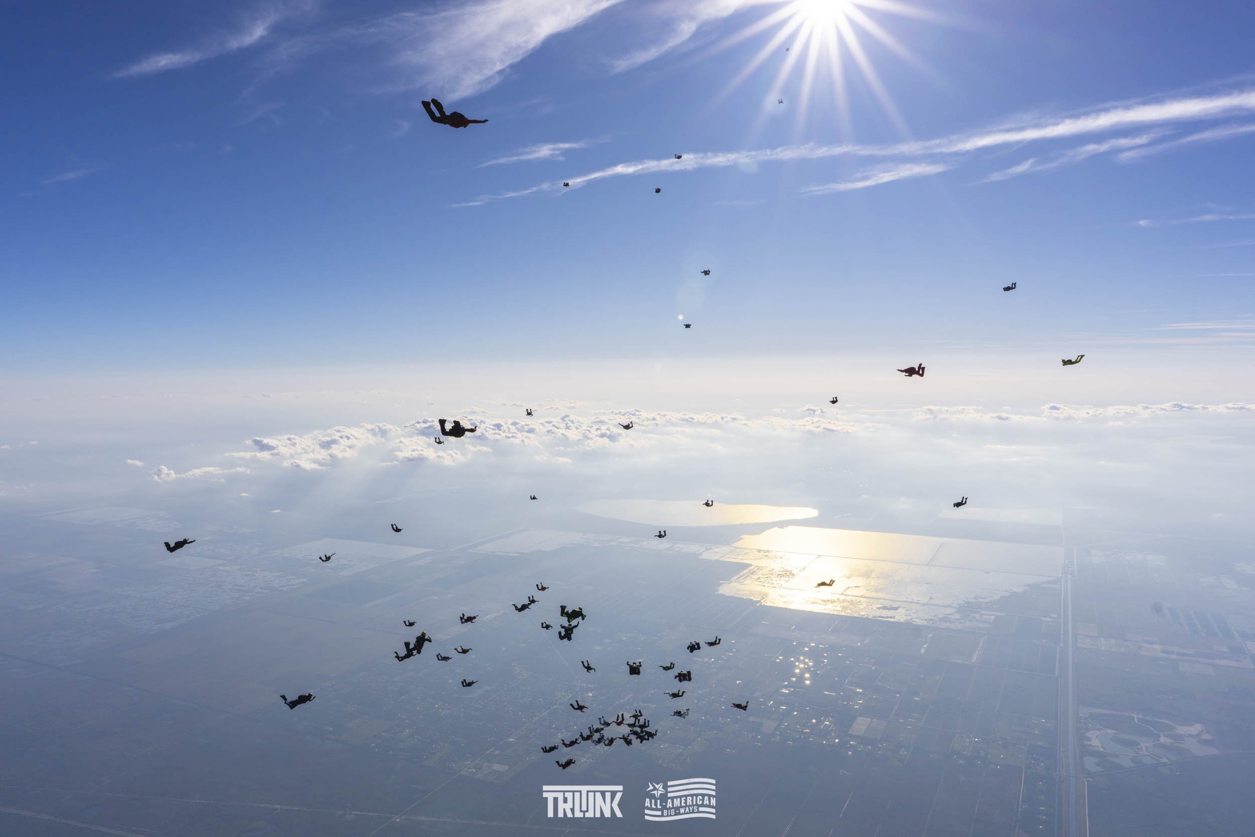 A group of skydivers free-falling from a plane over a body of water and farmland, with the sun shining brightly in a clear blue sky.