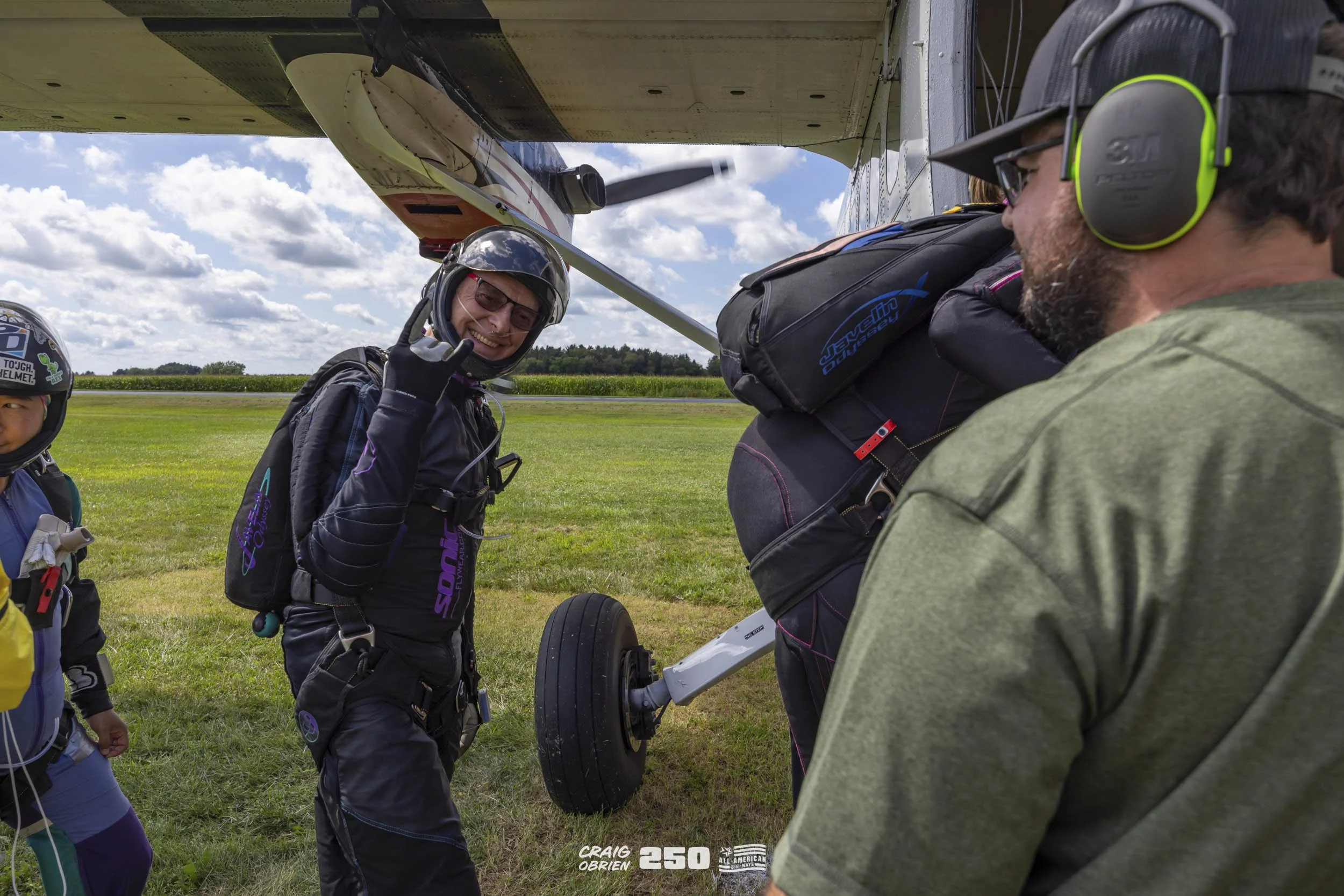A skydiver preparing to jump out of an aircraft, giving a thumbs-up and smiling, with a man wearing green ear protection and a gray hat standing nearby.