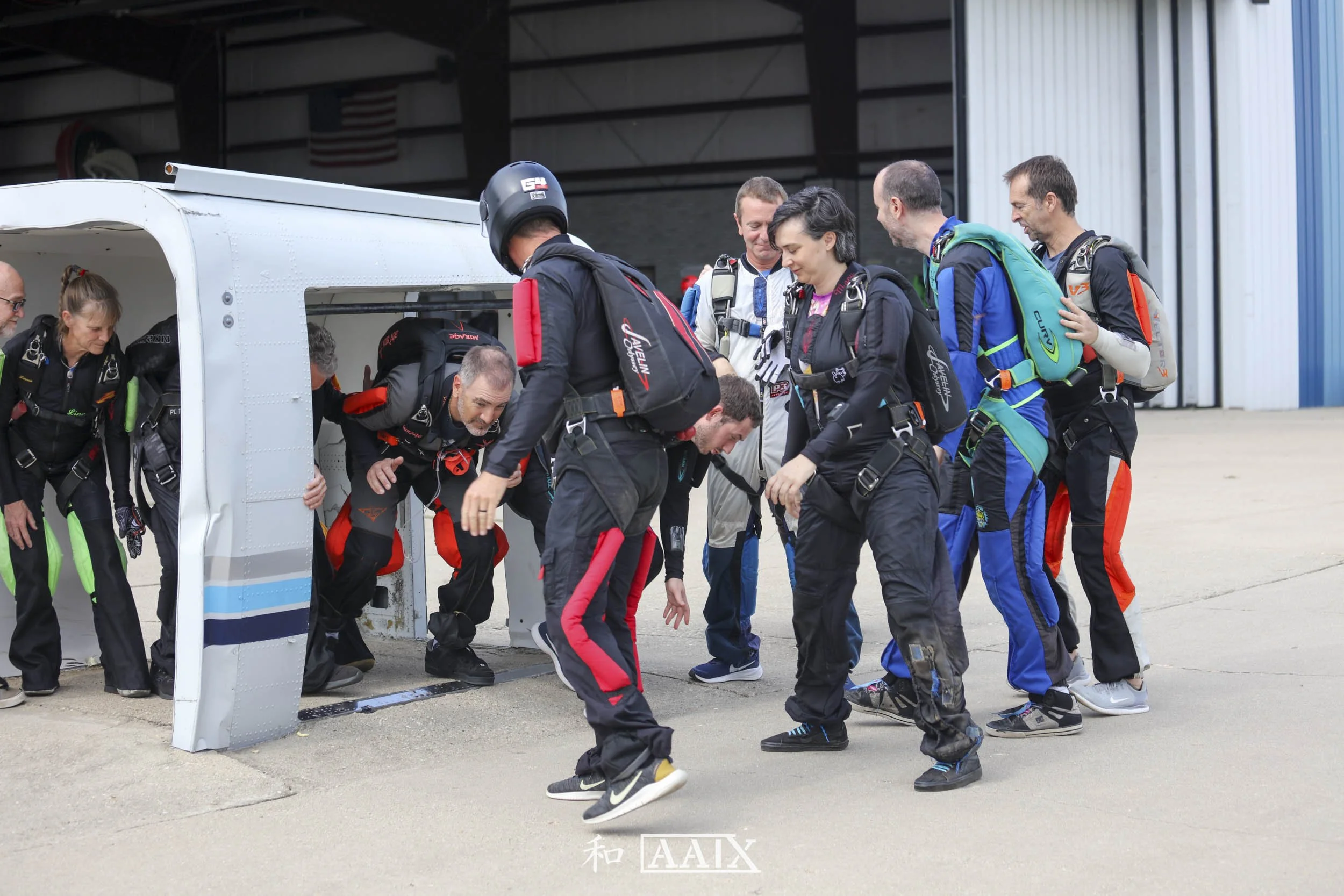 A group of skydivers in jumpsuits and harnesses preparing to exit an aircraft, with some outside the aircraft and others just inside, on an airport tarmac.