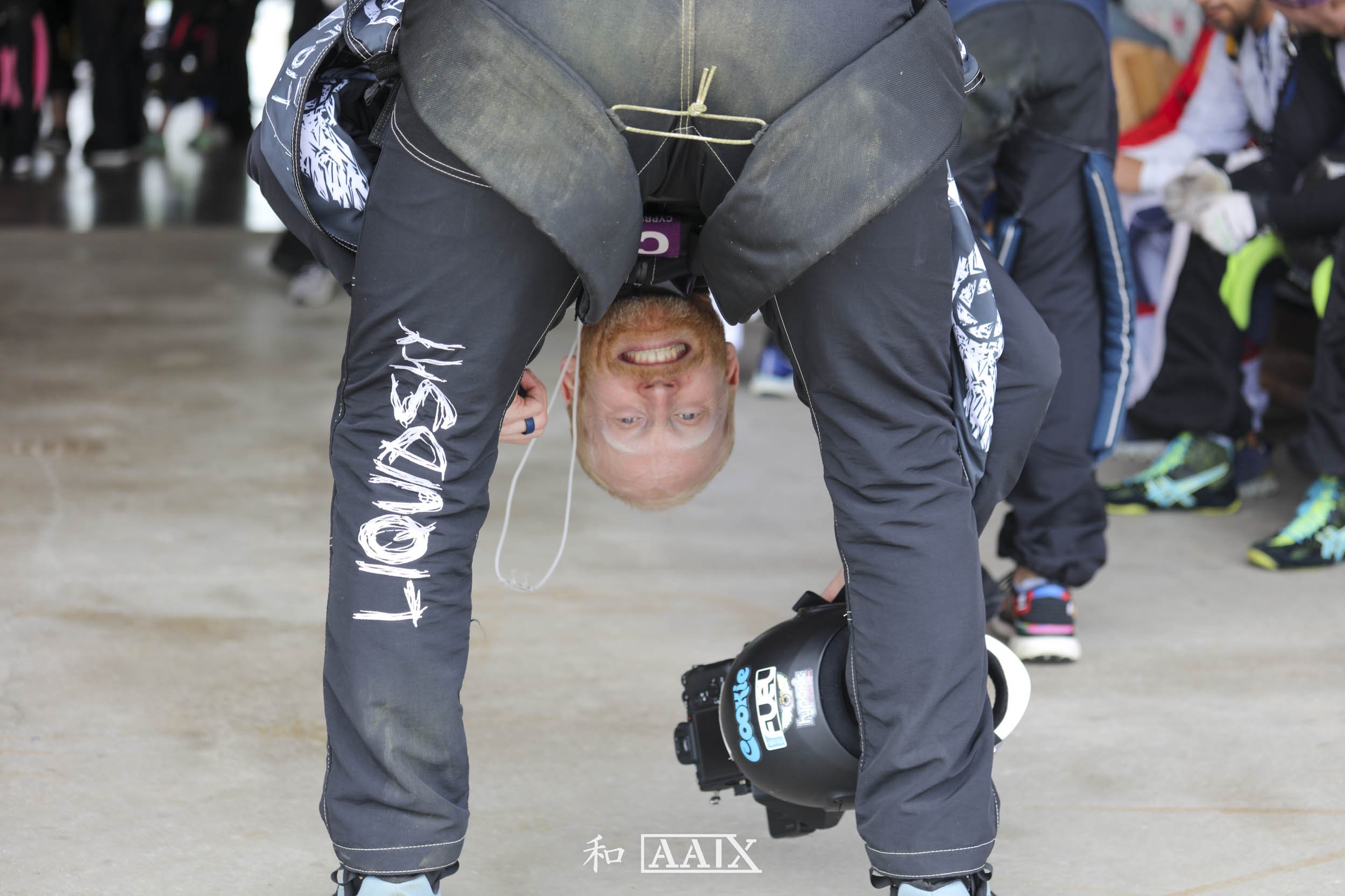 Man dressed in motorcycle racing gear bending forward with his head upside down, smiling, holding a helmet, standing on a concrete floor surrounded by other people.