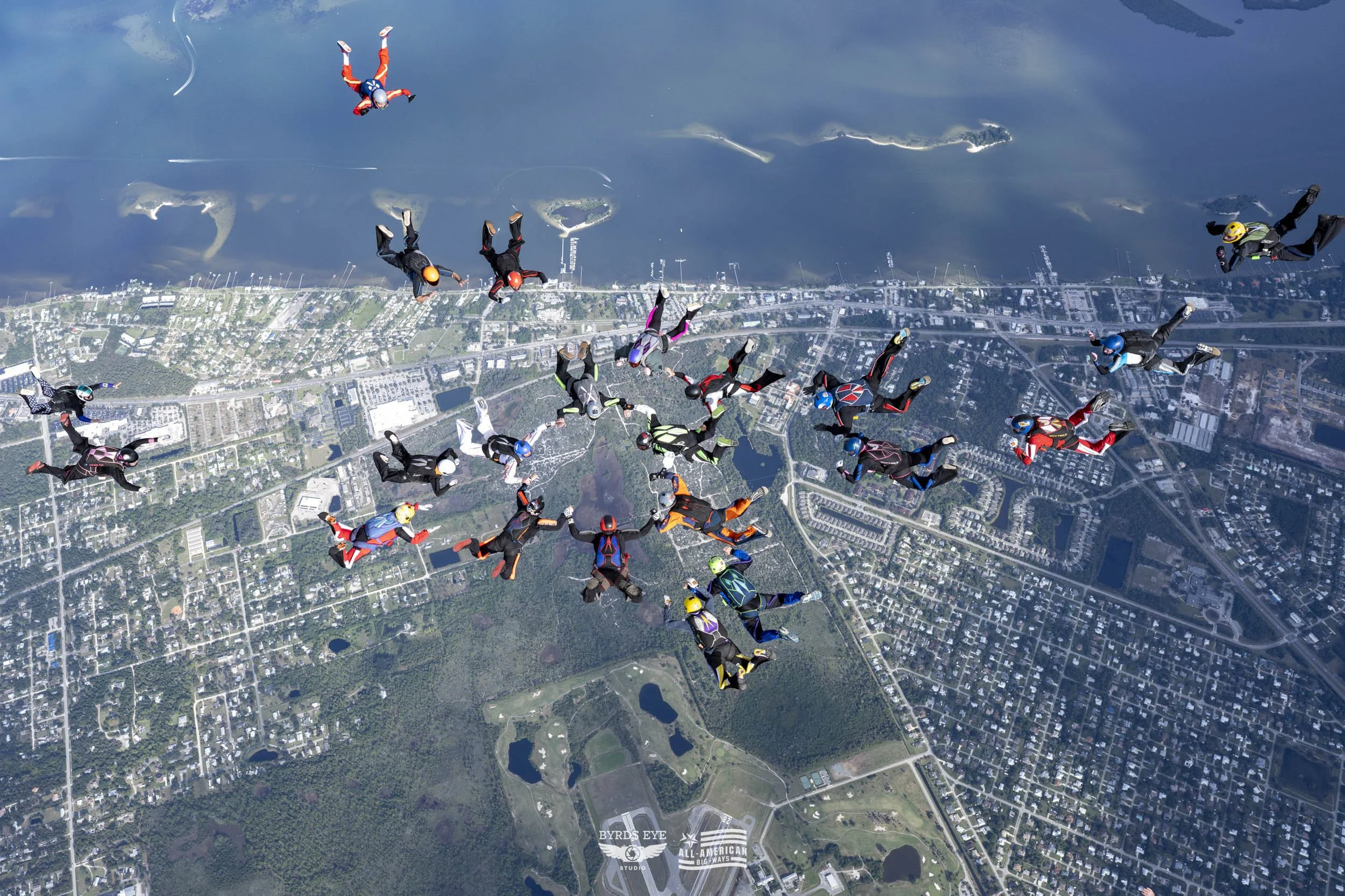 A large group of skydivers in colorful jumpsuits and helmets free-falling over a city landscape with lakes and roads.