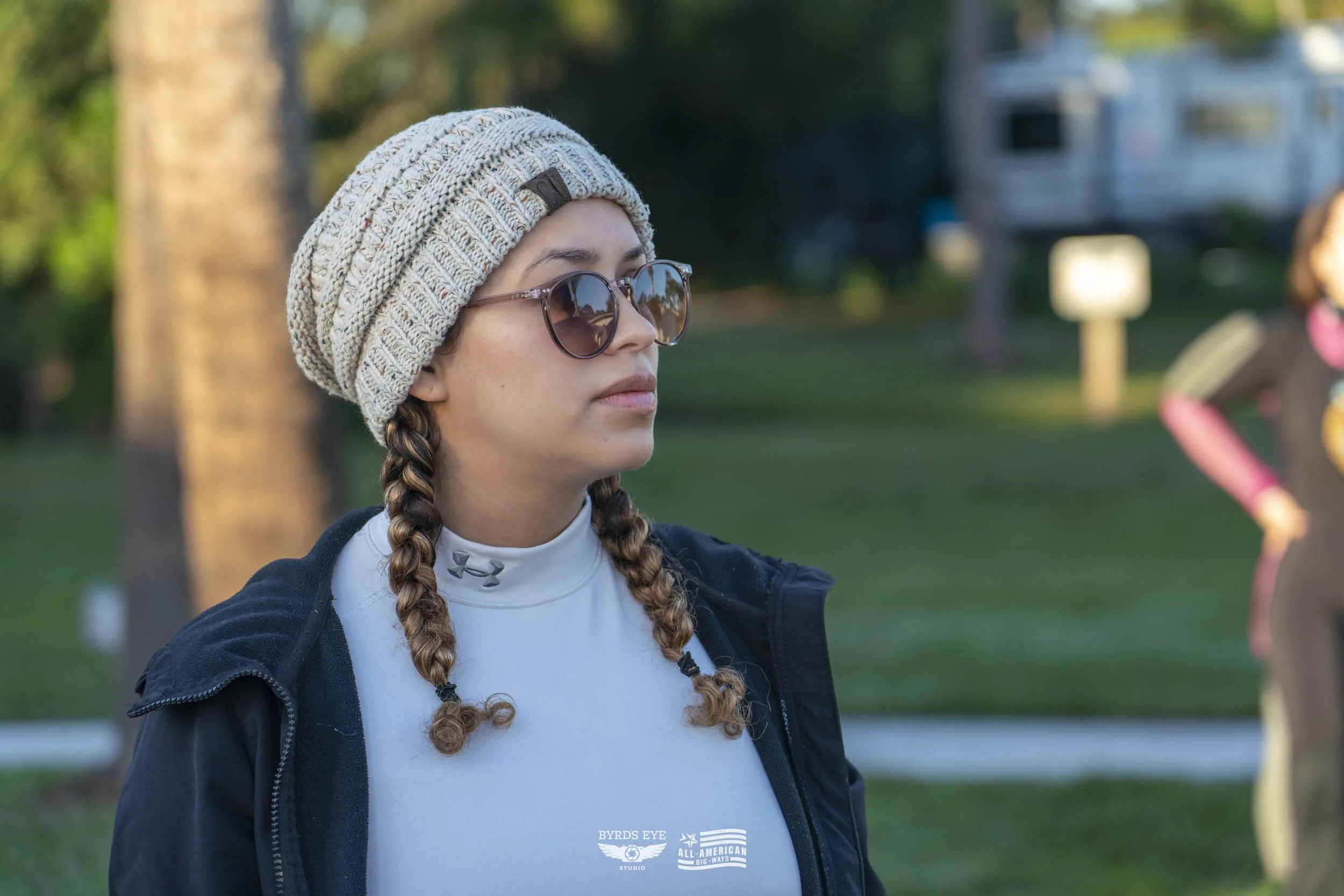 Young woman with braided hair, wearing sunglasses, a knit beanie, and sportswear, standing outdoors with a blurred background of trees, grass, and a person in the background.