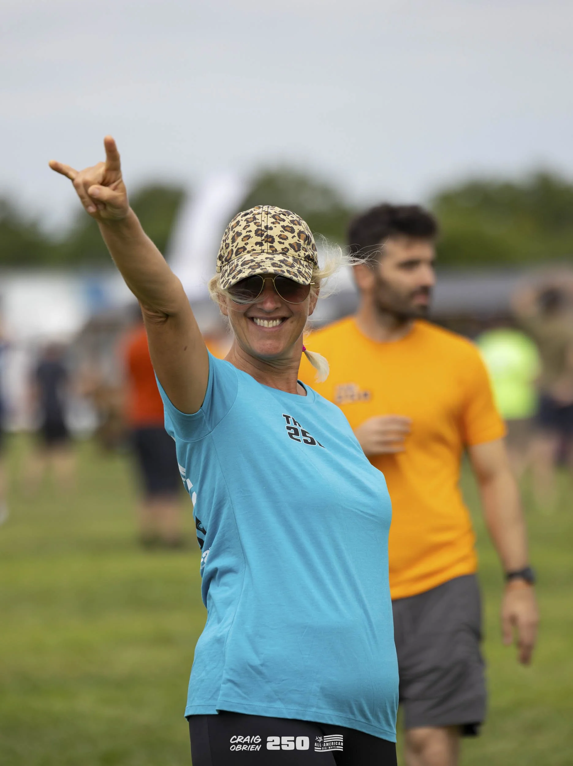 Woman smiling and making a rock and roll hand gesture at an outdoor event, wearing sunglasses, a leopard print cap, and a bright blue T-shirt, with a man in an orange T-shirt in the background.