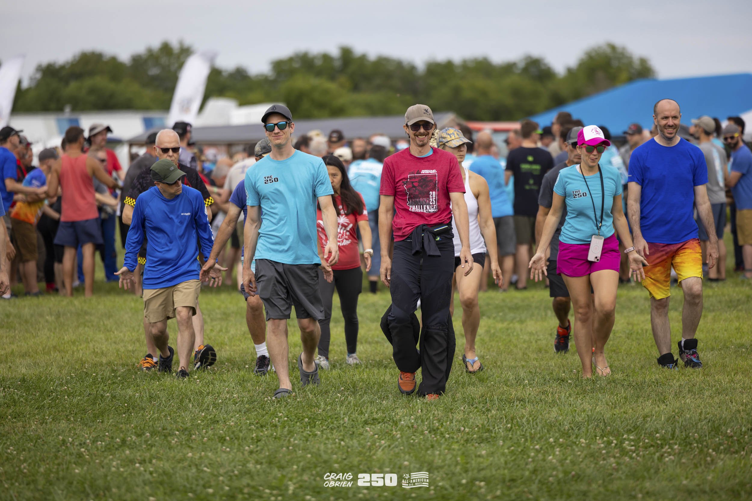 Group of people walking on a grassy field at an outdoor event, with tents and trees in the background, some wearing athletic clothing and accessories.