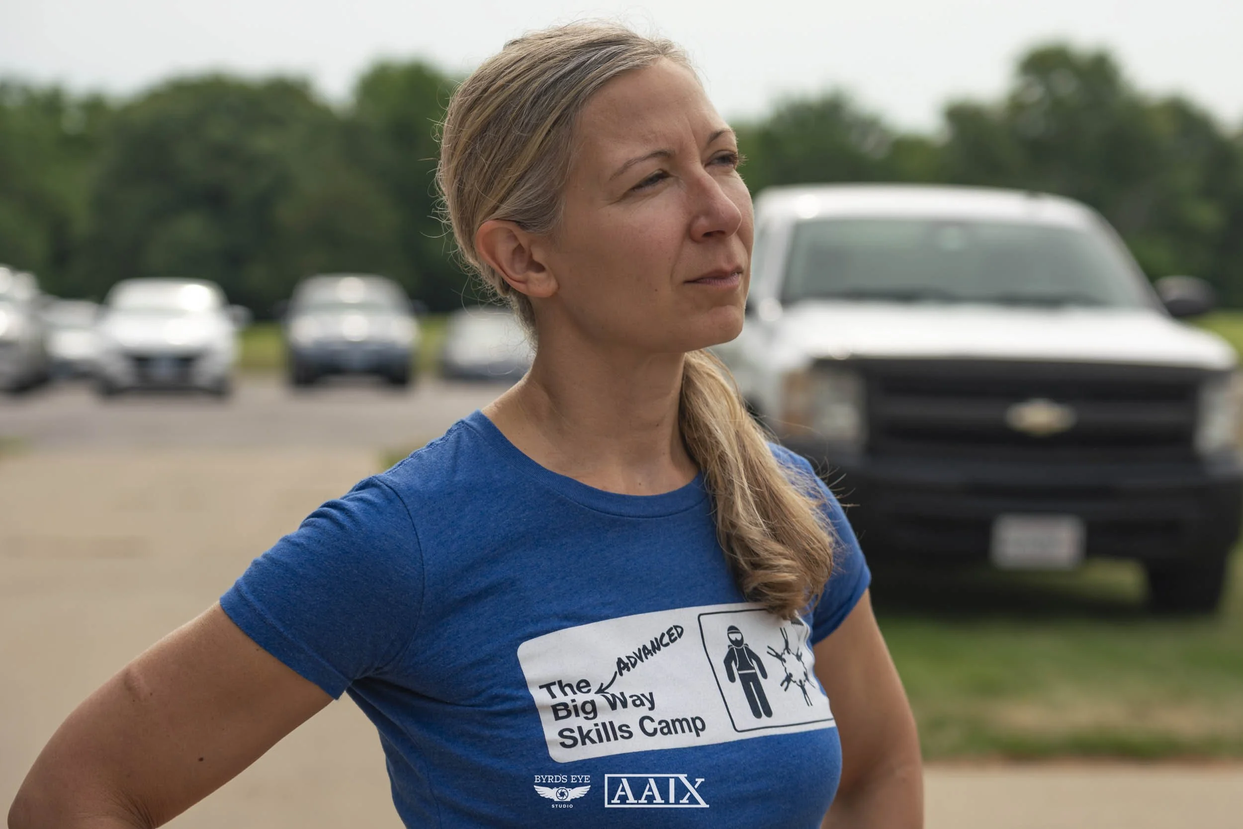 A woman with blonde hair tied back stands outdoors with a serious expression, wearing a blue T-shirt that reads 'The Advanced Big Way Skills Camp' and has graphics related to skydiving.