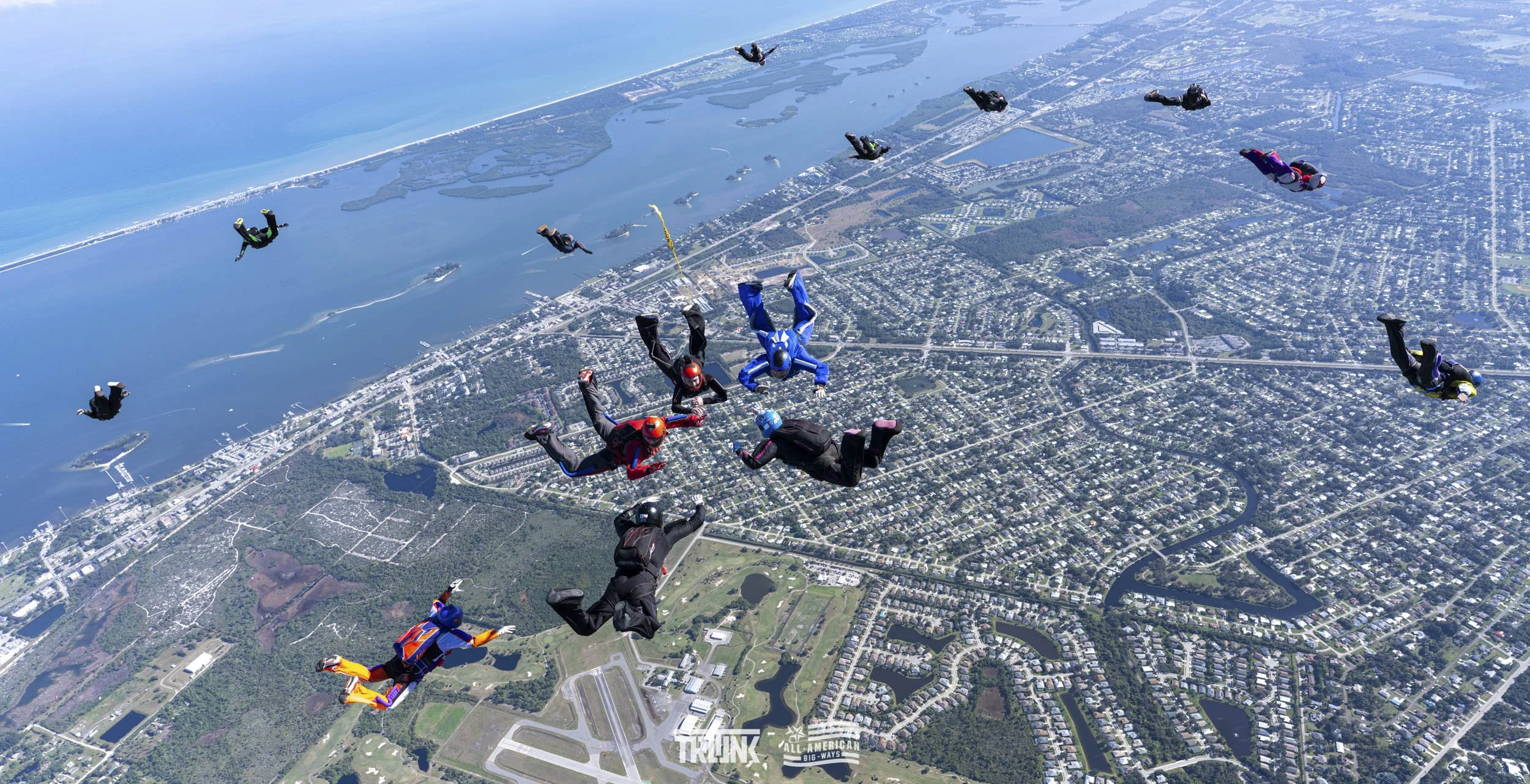 Multiple skydivers in freefall over a city, with water and land visible below.
