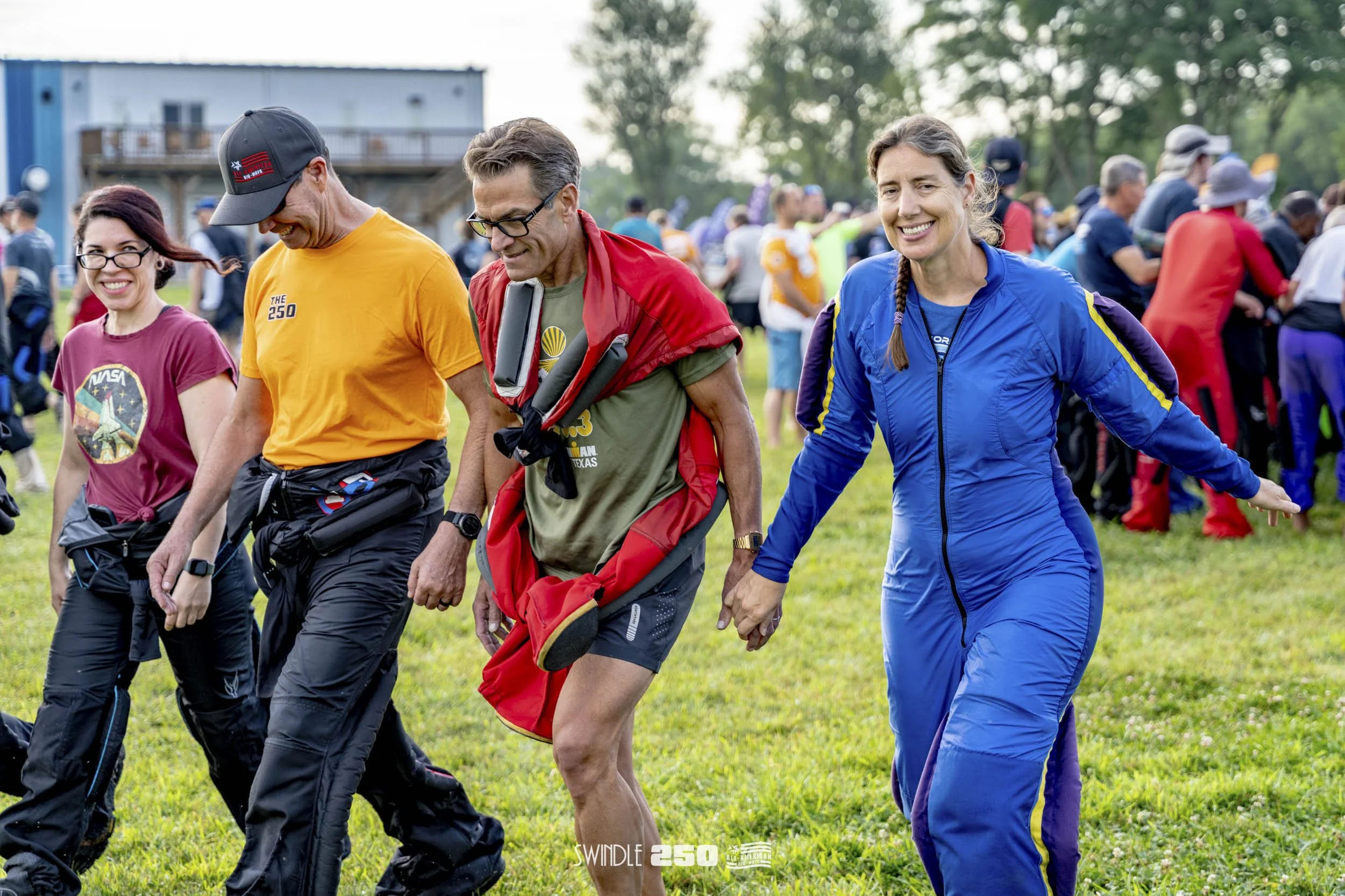 A group of people walking together outdoors during a daytime event, with a woman in a blue jumpsuit leading, and others smiling and holding hands.