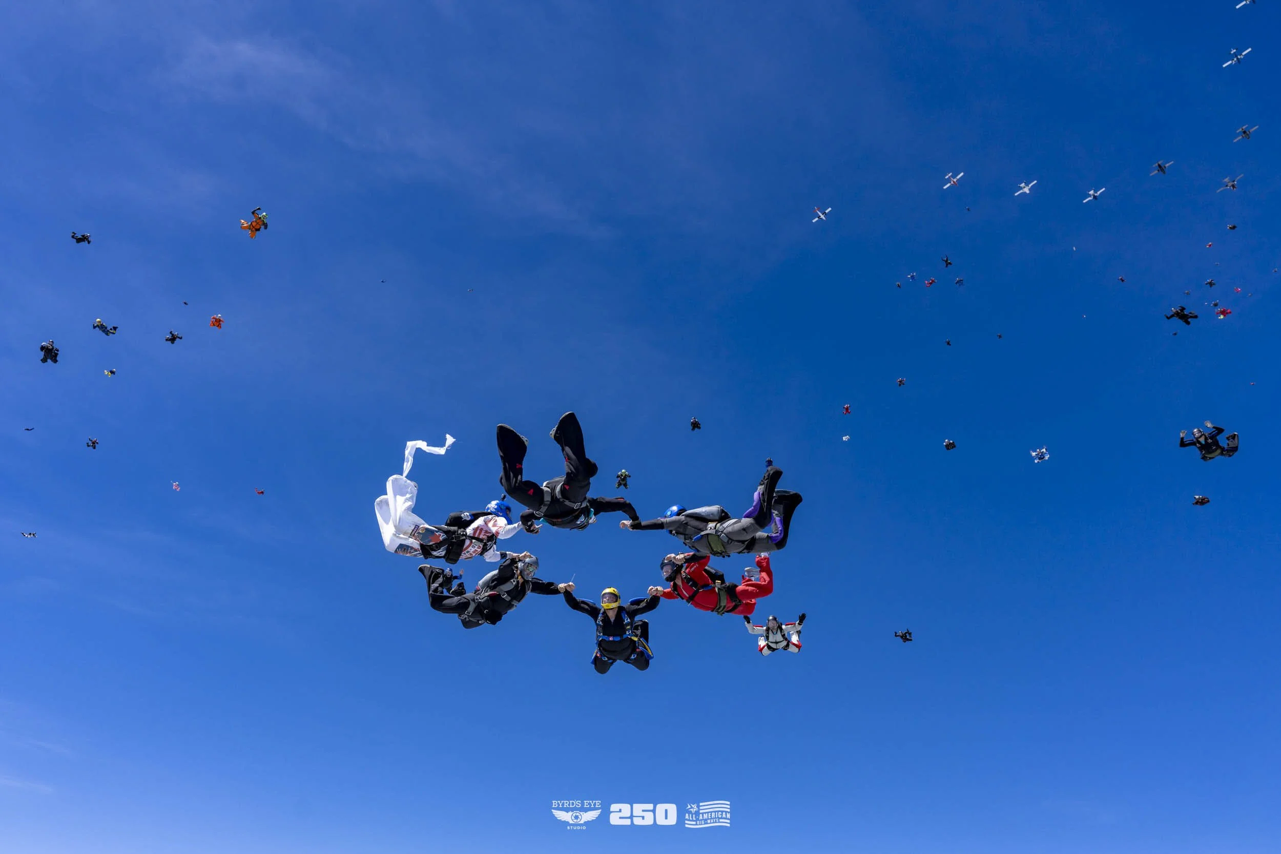 A group of skydivers holding hands in freefall with a blue sky and numerous other skydivers in the background. The bottom of the image features logos and the number '250'.
