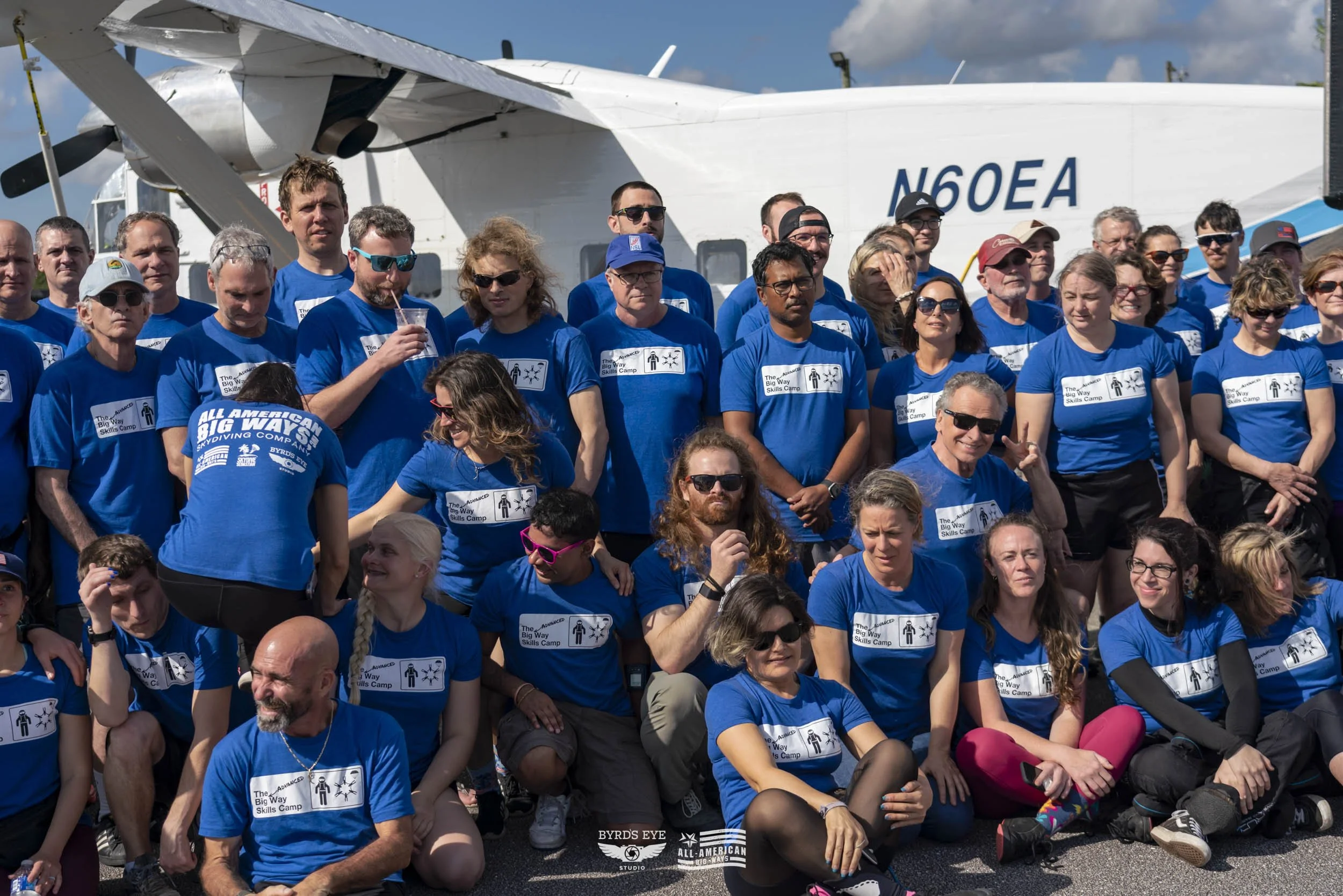 Group of people in blue T-shirts gathered outdoors in front of a large airplane, some sitting and some standing, during a sunny day.