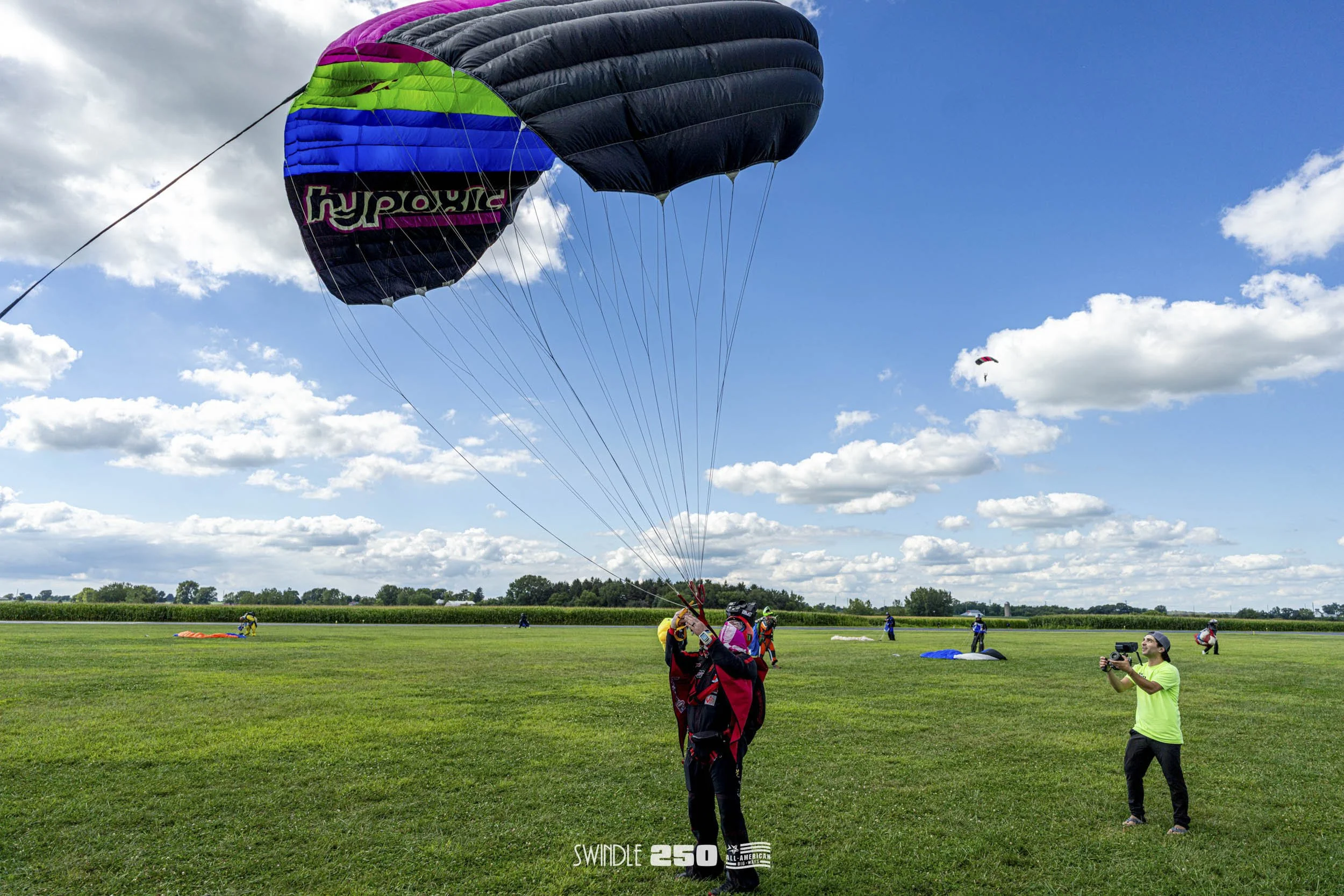 Person preparing for a tandem skydive with a black and purple parachute, standing on a grassy field, with a photographer recording the scene and other parachutes on the ground in the background.