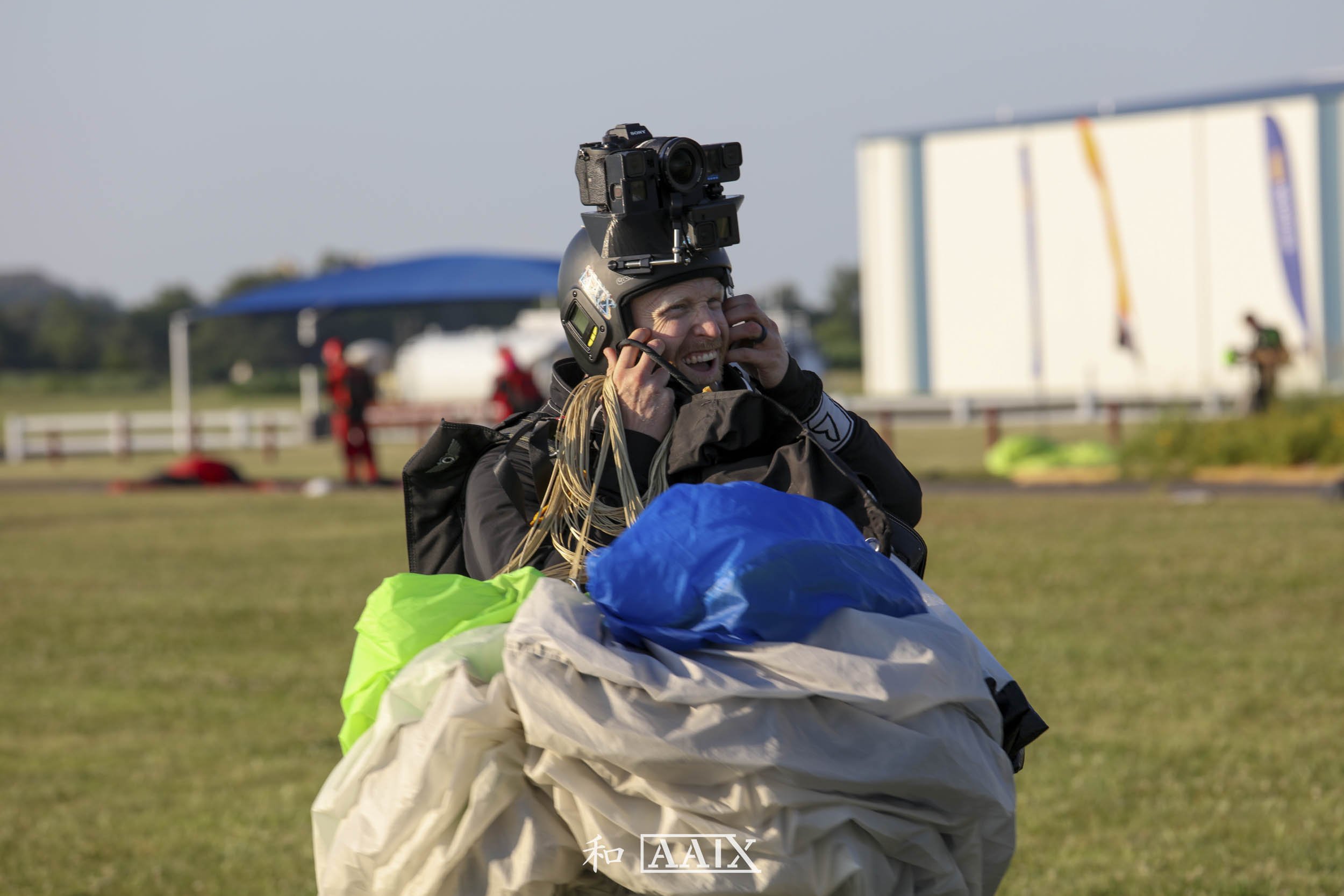 A skydiver wearing a helmet with a camera, holding a parachute, smiling on a grassy field with other skydivers and equipment in the background.