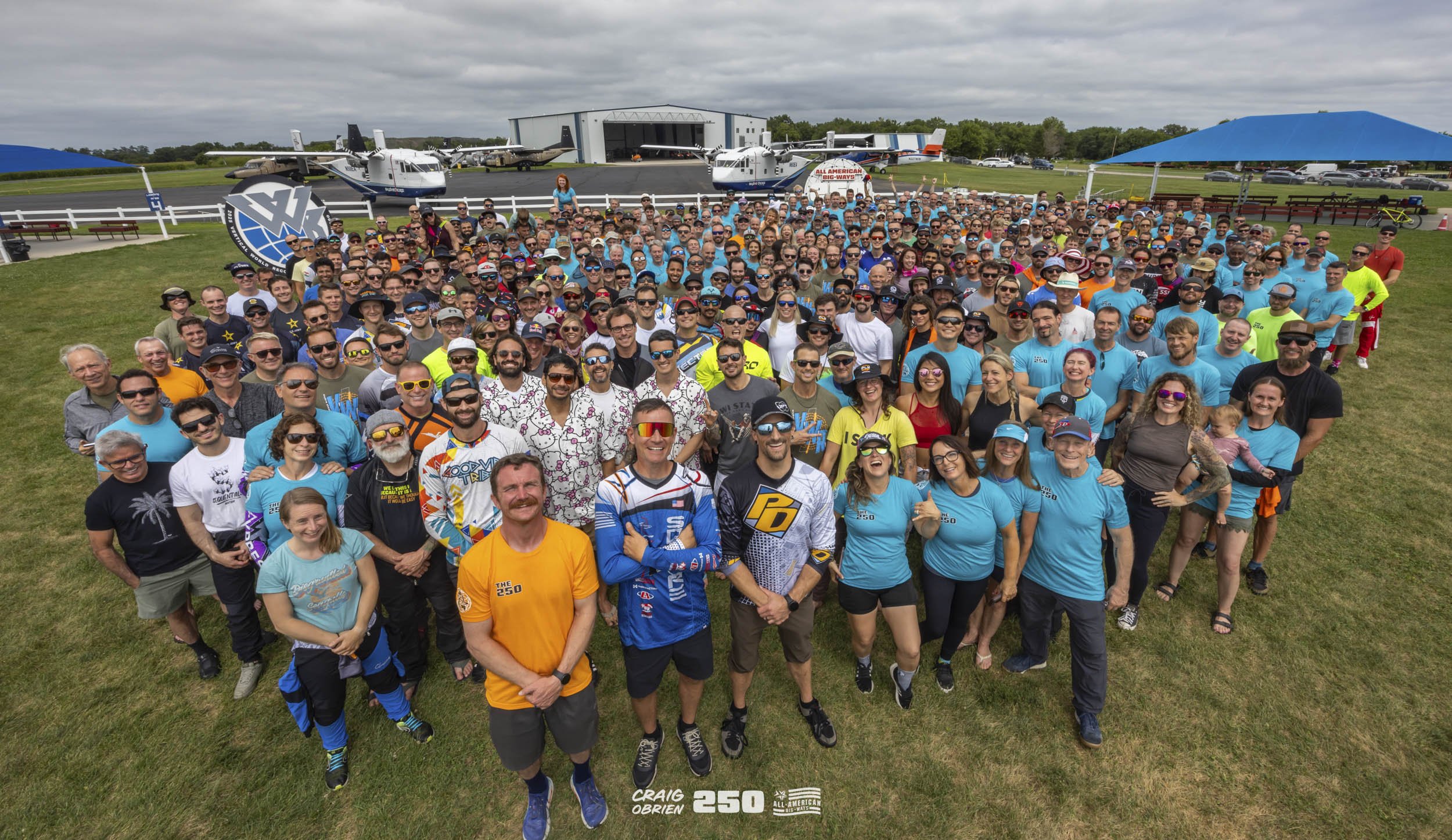 A large group of people gathered on grass at an airfield with small airplanes and hangar in the background. Many are wearing promotional or event-branded clothing, with some wearing sunglasses and hats. The gathering appears to celebrate an aviation 