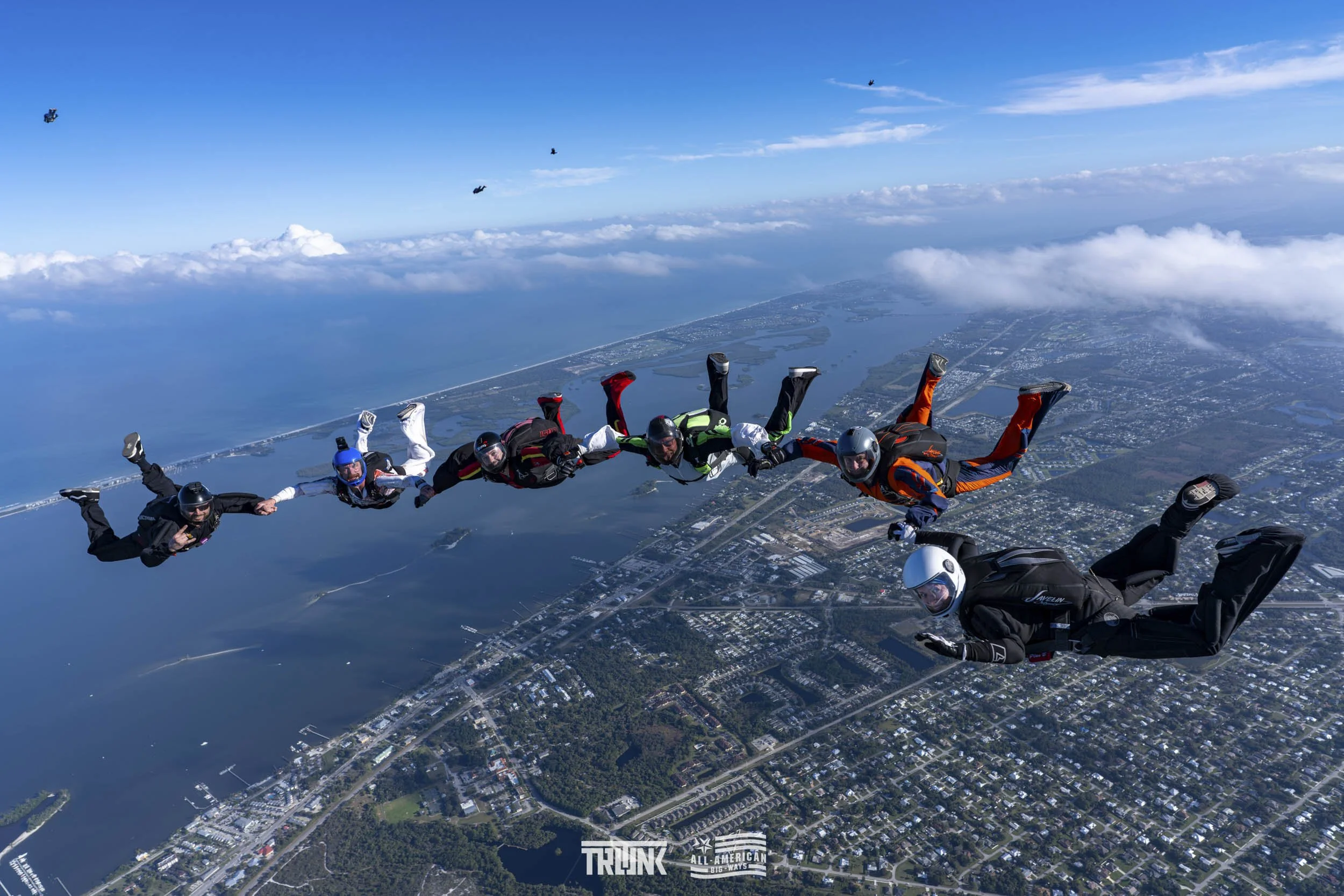 Skydivers in free fall formation over a city with bodies of water and clouds in the background.