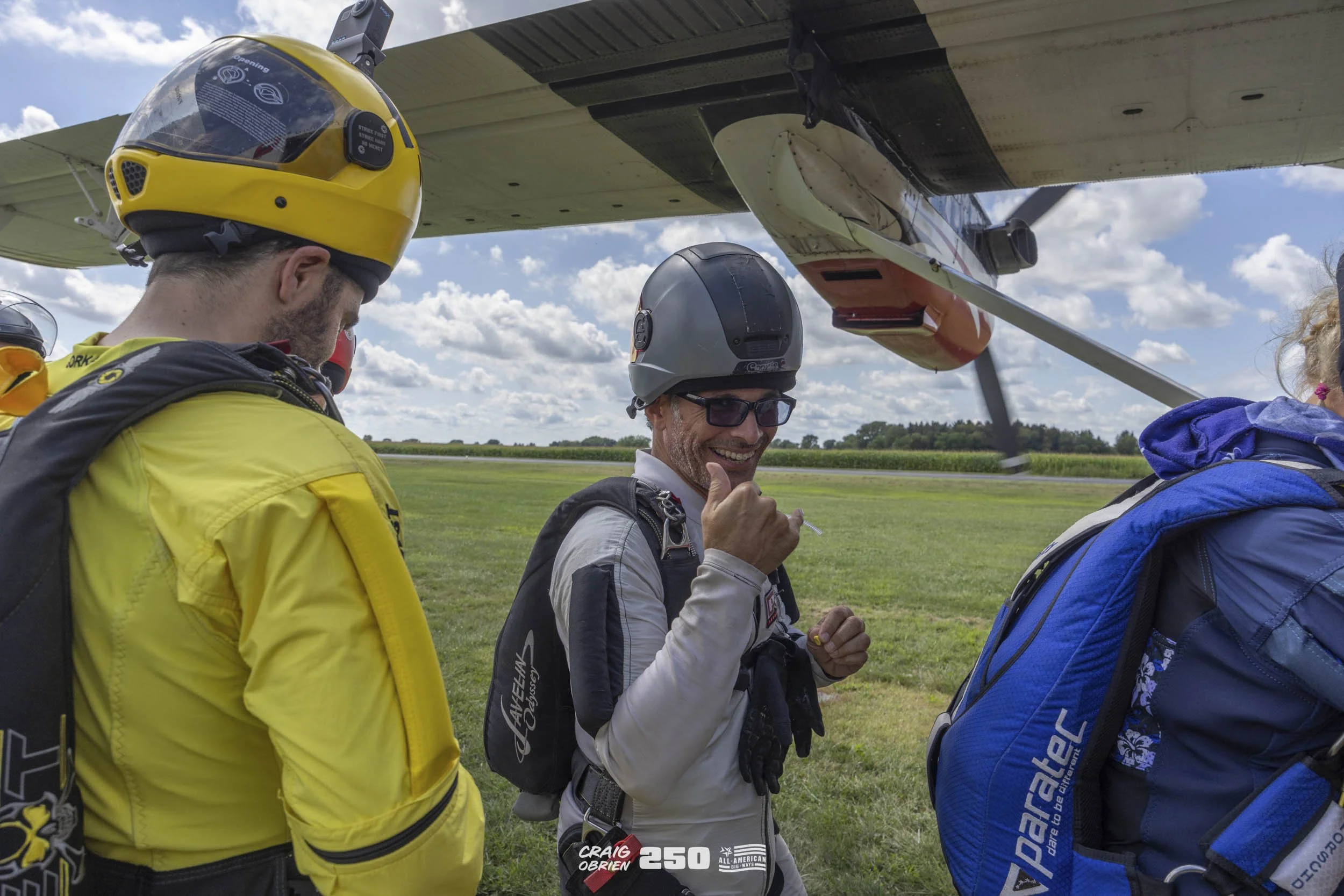 Three skydivers in jumpsuits and helmets stand on grass near an airplane, preparing for a jump. One of them is smiling and giving a thumbs-up.