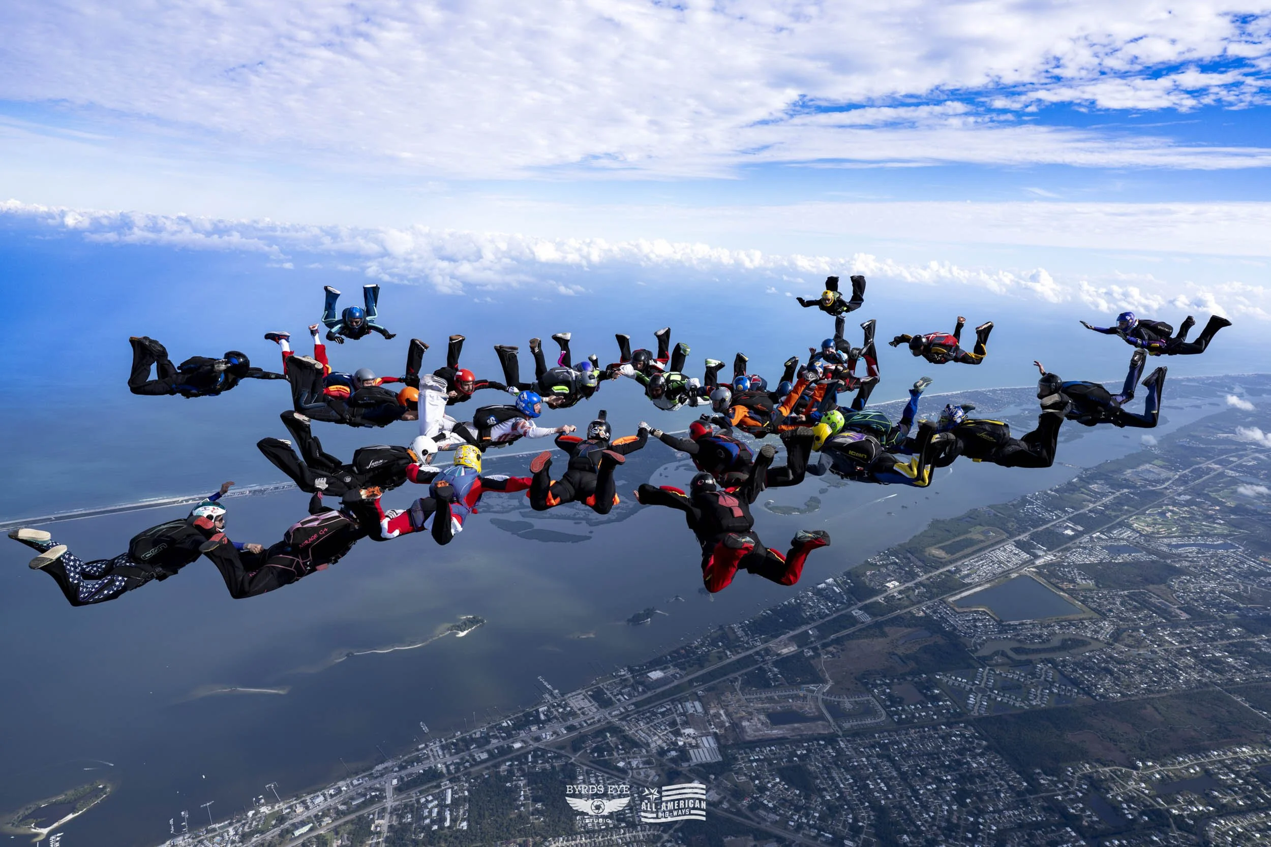 Group of skydivers in freefall formation over a landscape with water and city roads below.