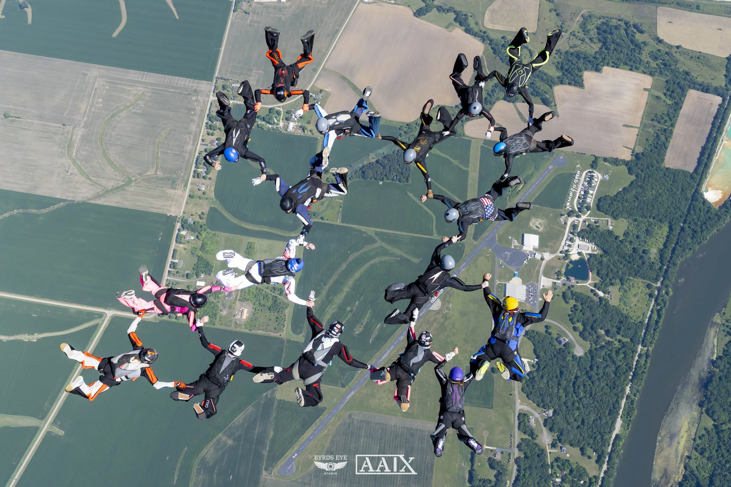 Skydivers forming a circle in freefall over a rural landscape with fields, roads, and a river.