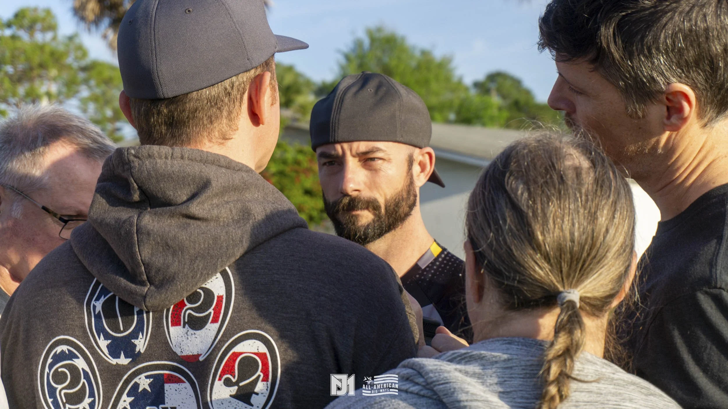 Group of people engaged in serious discussion outdoors, wearing casual and sports clothing, with trees and a house in the background.