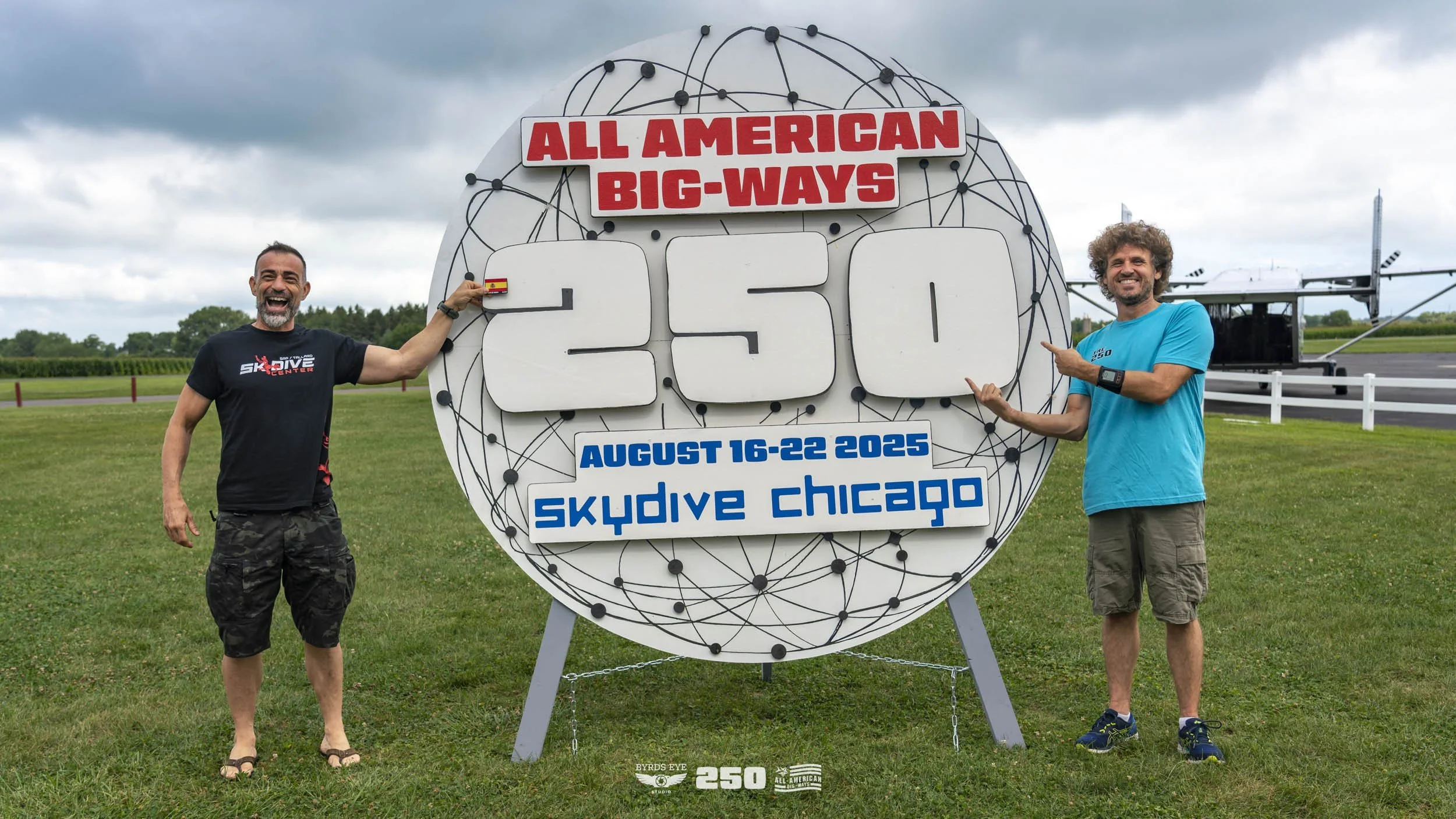 Two smiling men stand outdoors near a large sign that reads 'ALL AMERICAN BIG-WAYS 250 AUGUST 16-22 2025 skydive chicago'. One man is pointing at the sign, and the other is touching it. The man on the left has a beard, wears a black t-shirt and camou