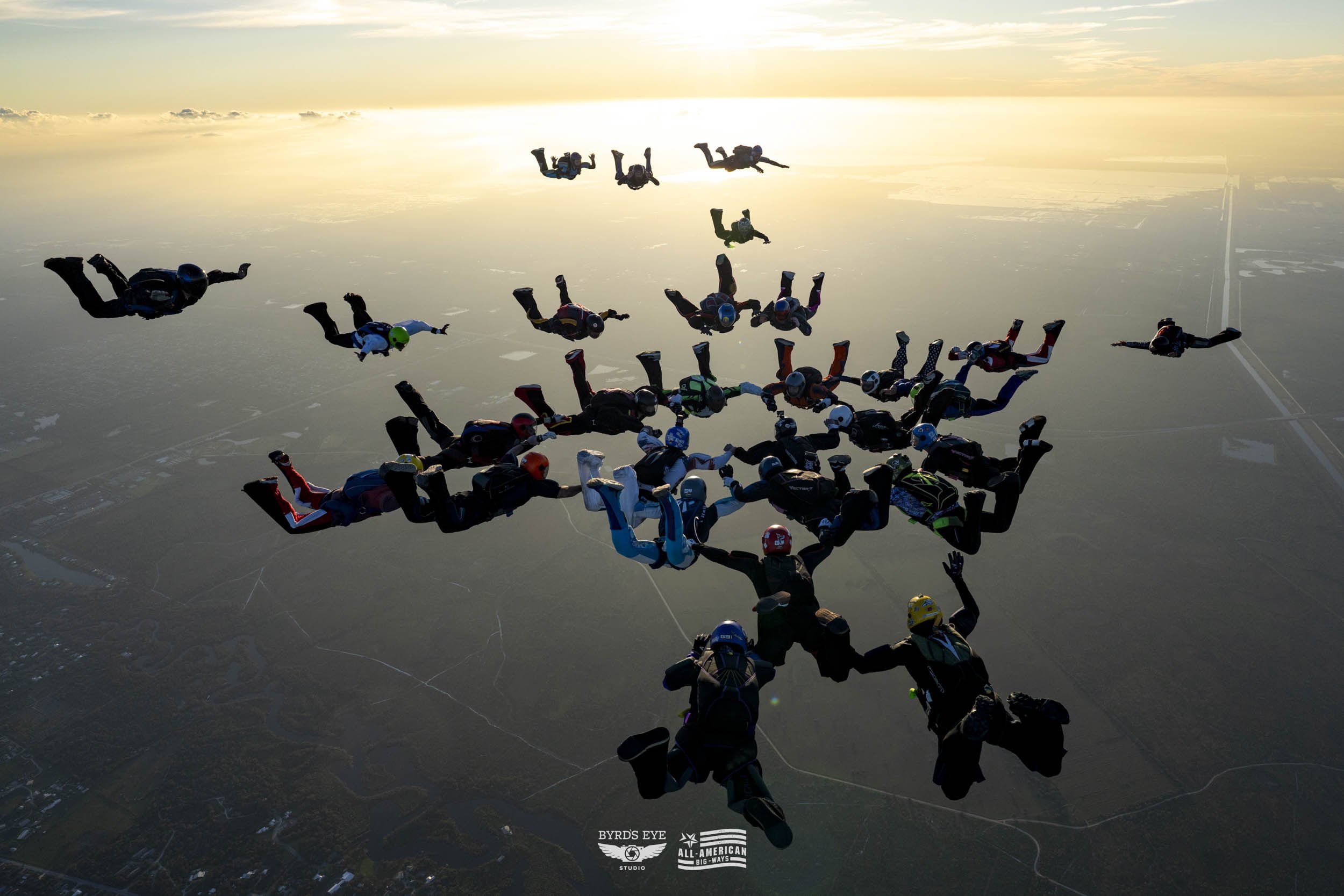 Group of skydivers forming a heart shape in freefall over a landscape during sunset.