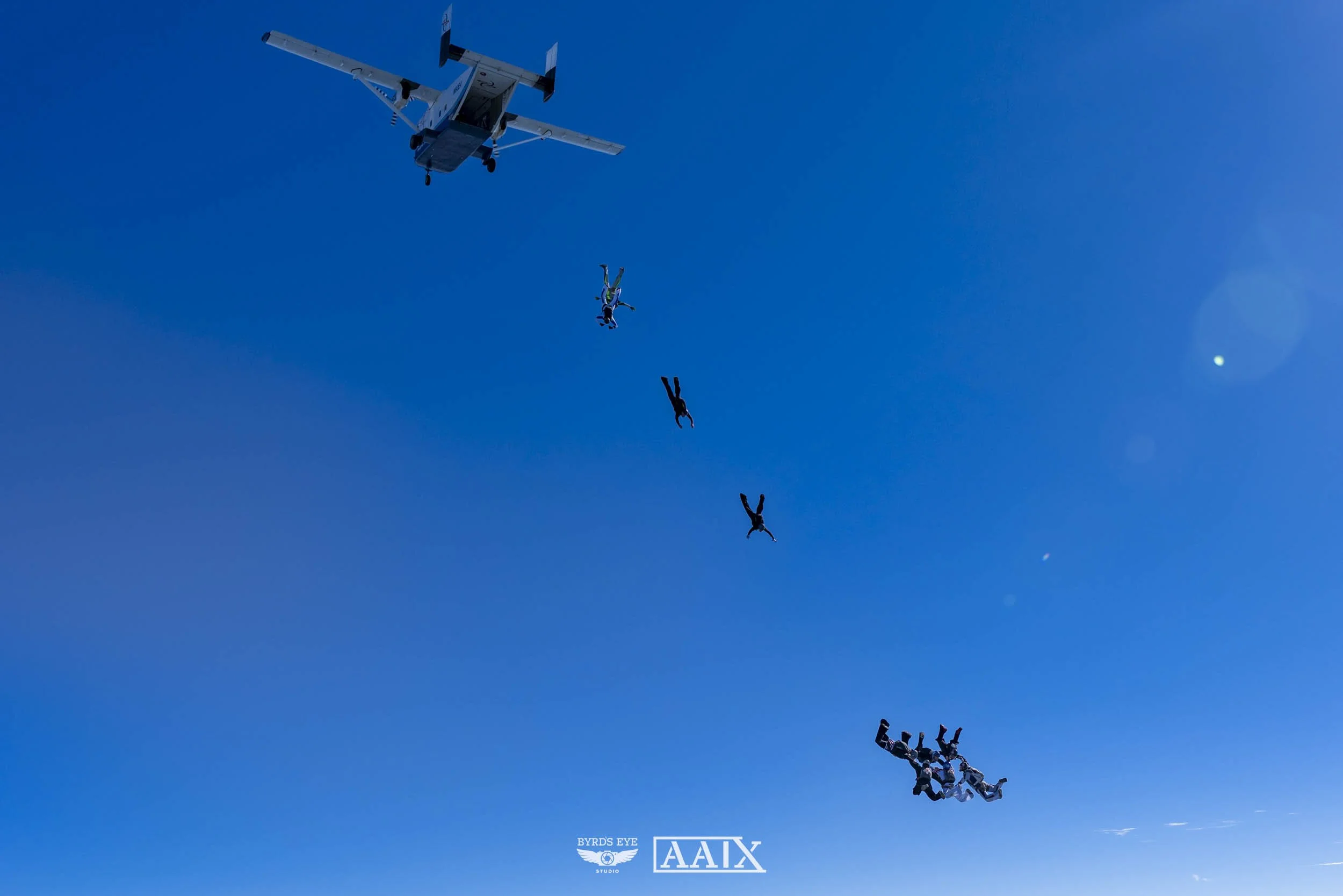 Skydivers mid-air during a jump, with an airplane visible above them against a clear blue sky.