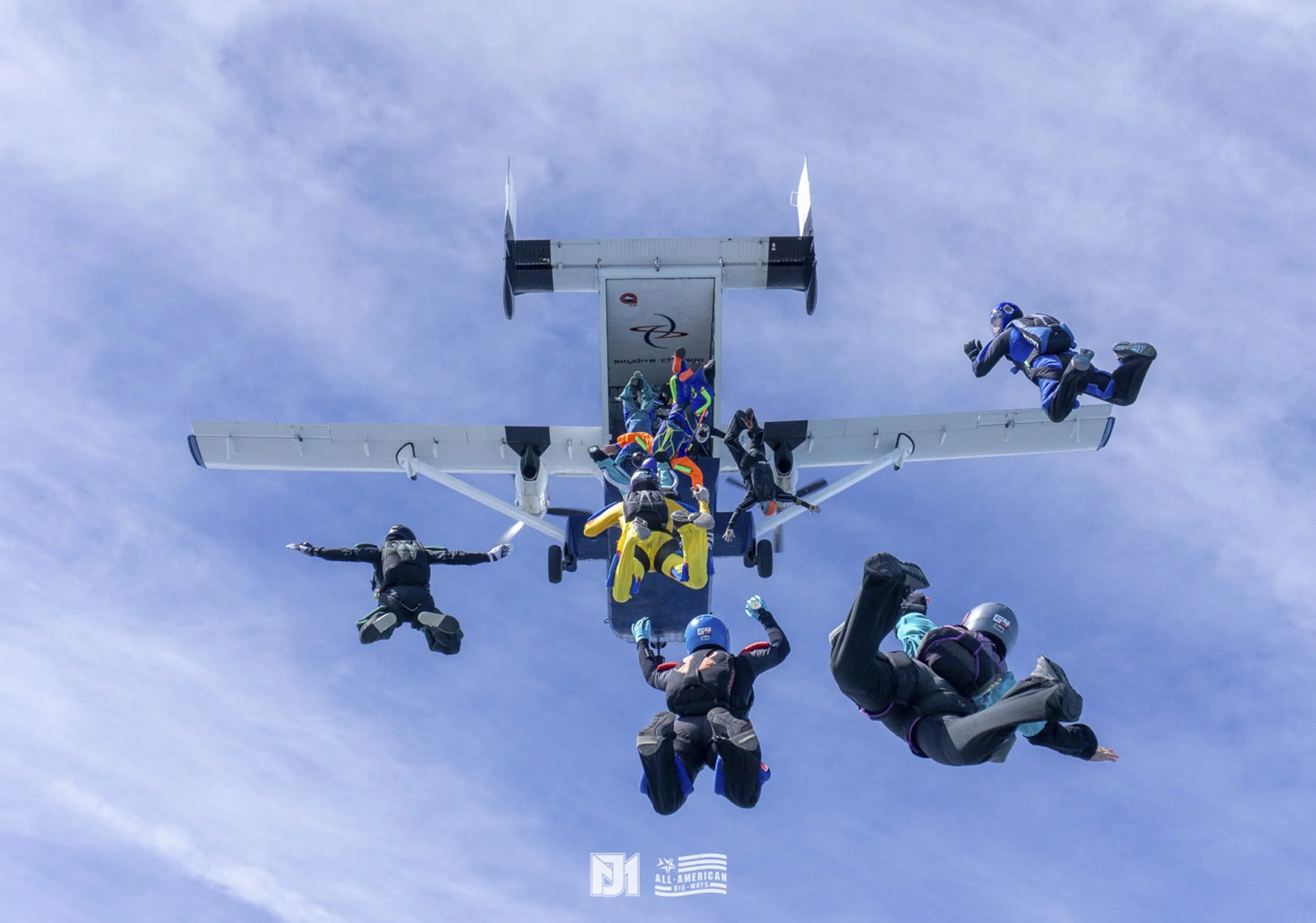 Skydivers mid-air after exiting an airplane, with blue sky and light clouds in the background.