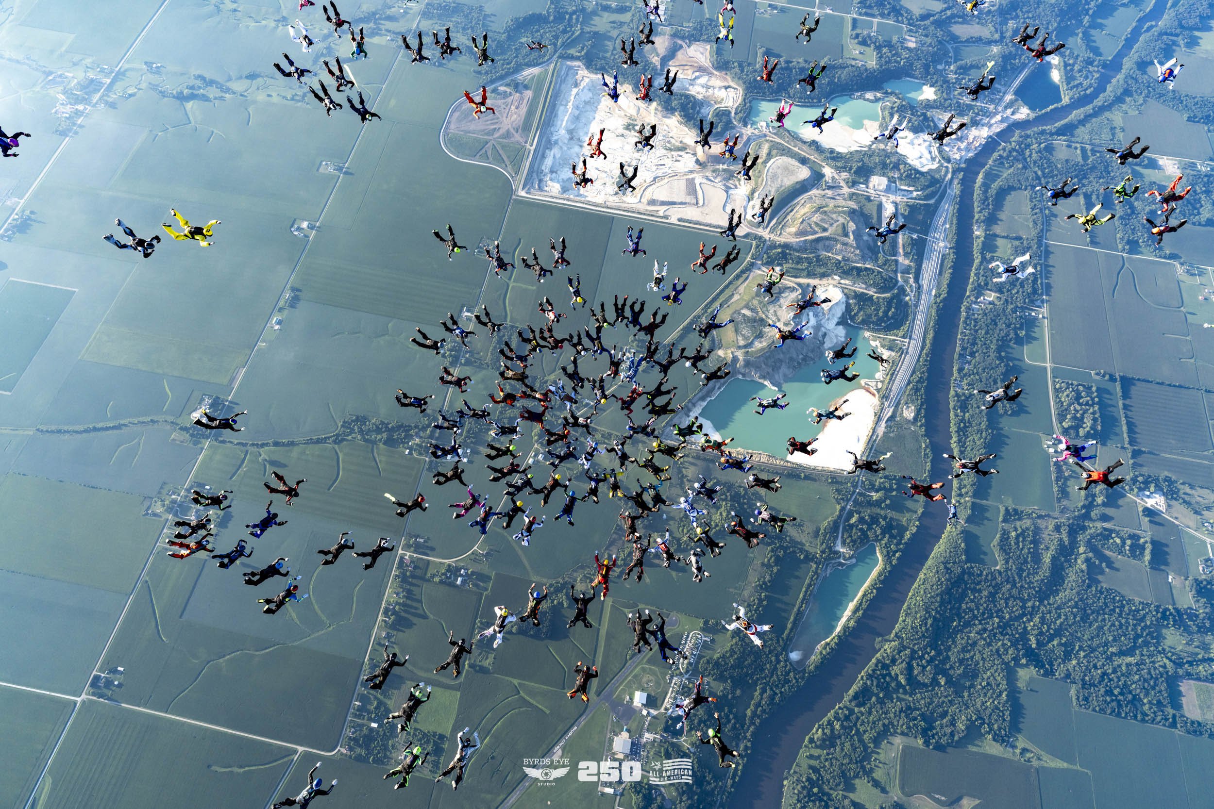 Group of skydivers in free fall above a landscape with fields, lakes, and forests.