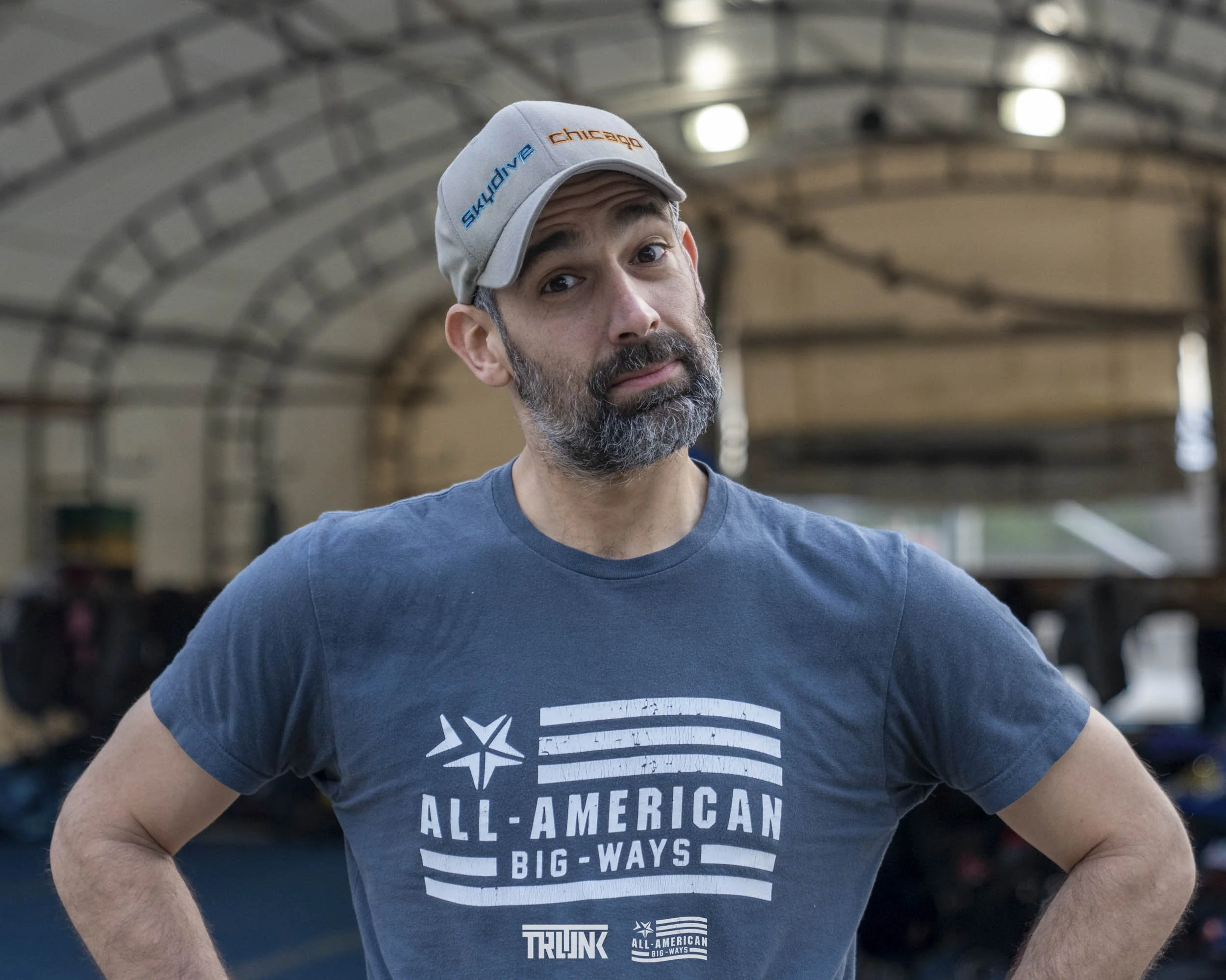 A man with a beard and mustache wearing a light gray baseball cap and a navy blue T-shirt standing with hands on hips inside a large indoor facility with a curved ceiling.