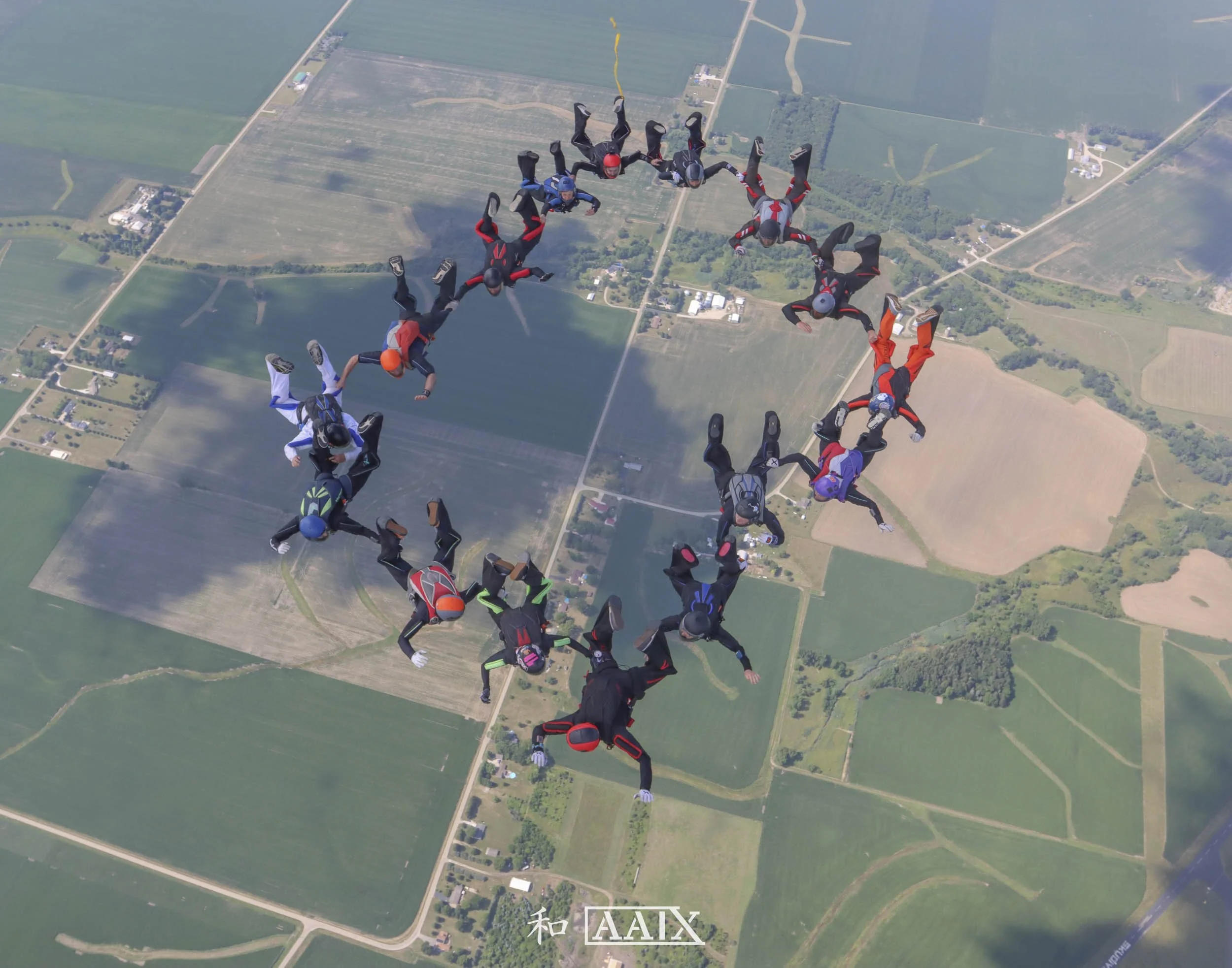 Group of skydivers forming a heart-shaped formation during free fall above a rural landscape with fields and roads.