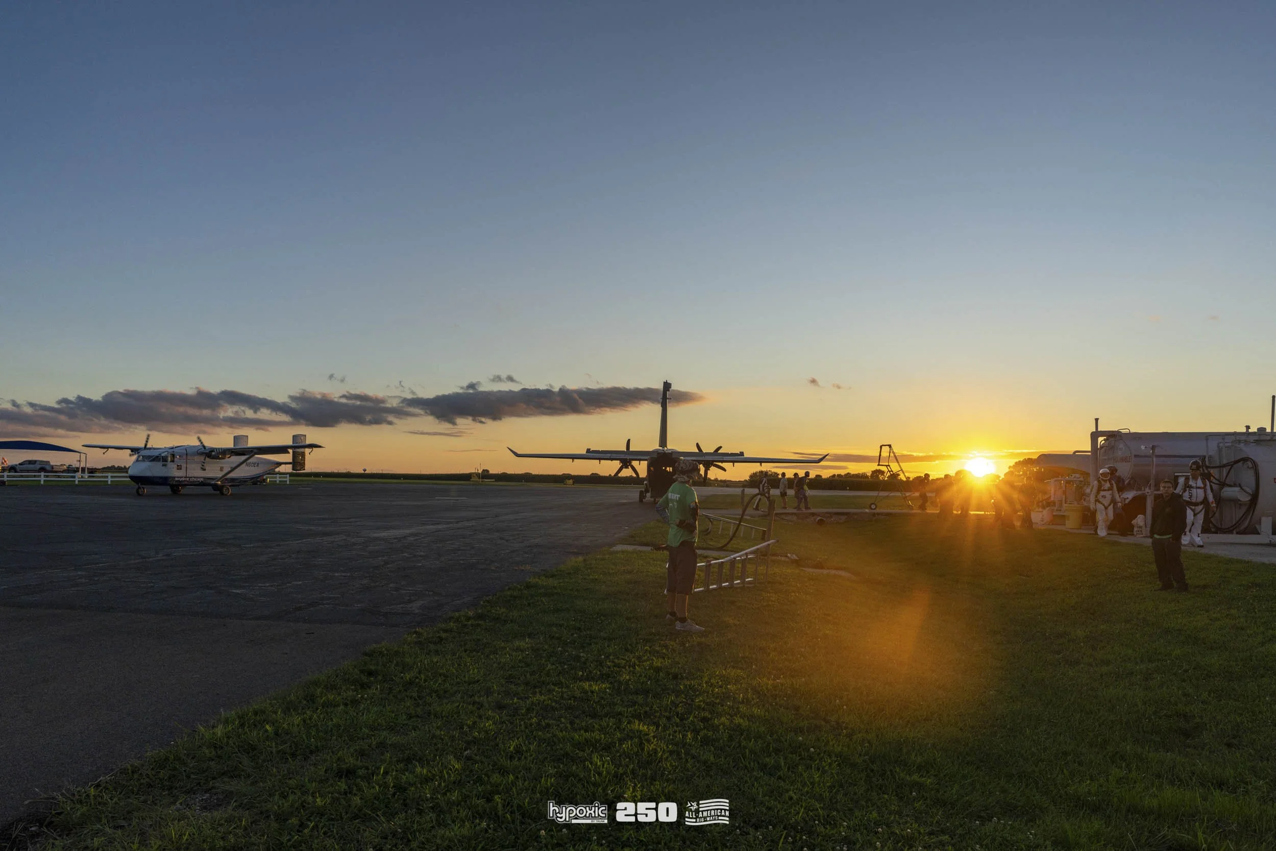 Sunset at an airfield with small aircraft parked on the tarmac, and people walking and working near the planes, with the sun near the horizon casting a warm glow.