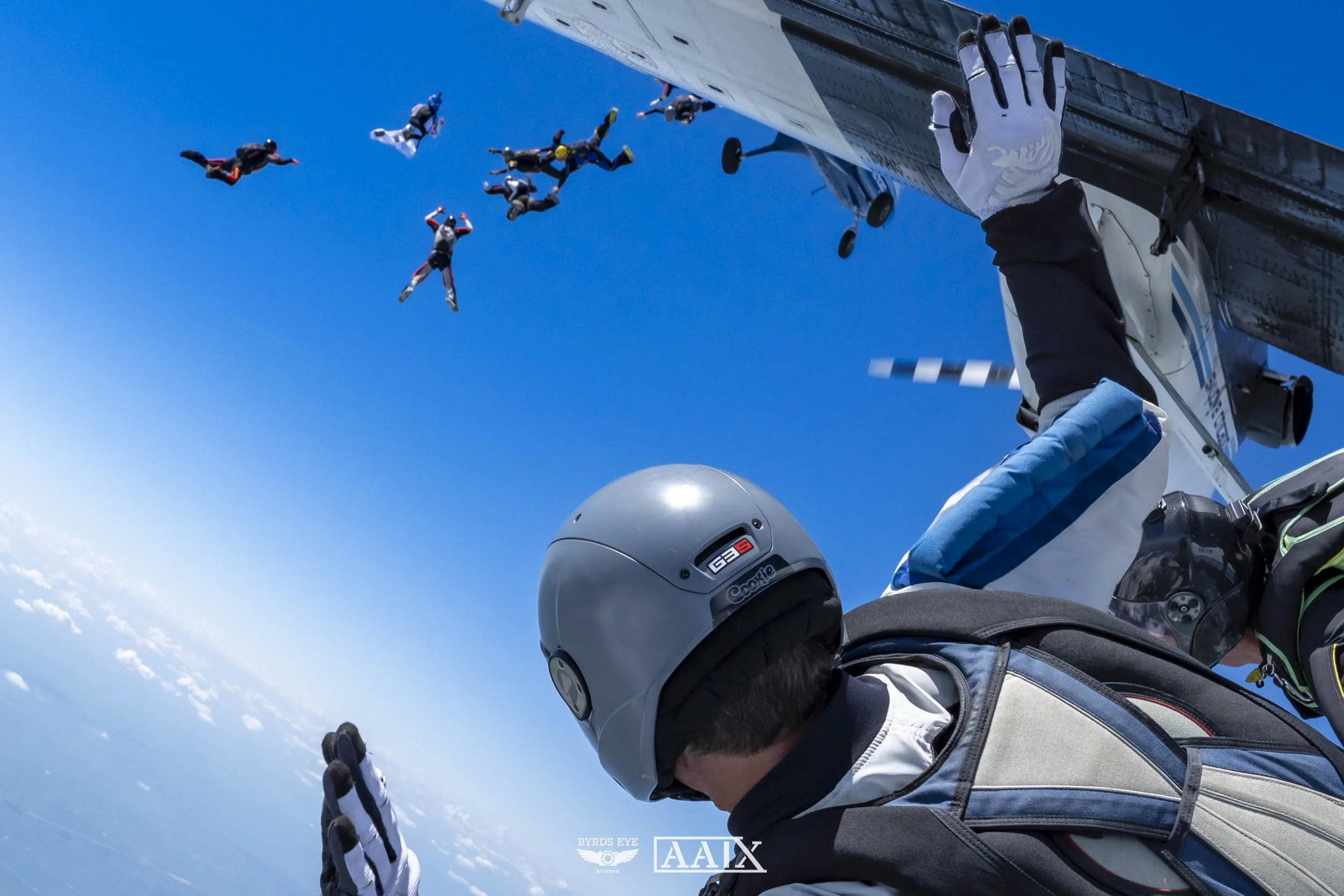 A skydiver is holding onto an aircraft door with one hand while skydiving with a group of divers in free fall against a blue sky.