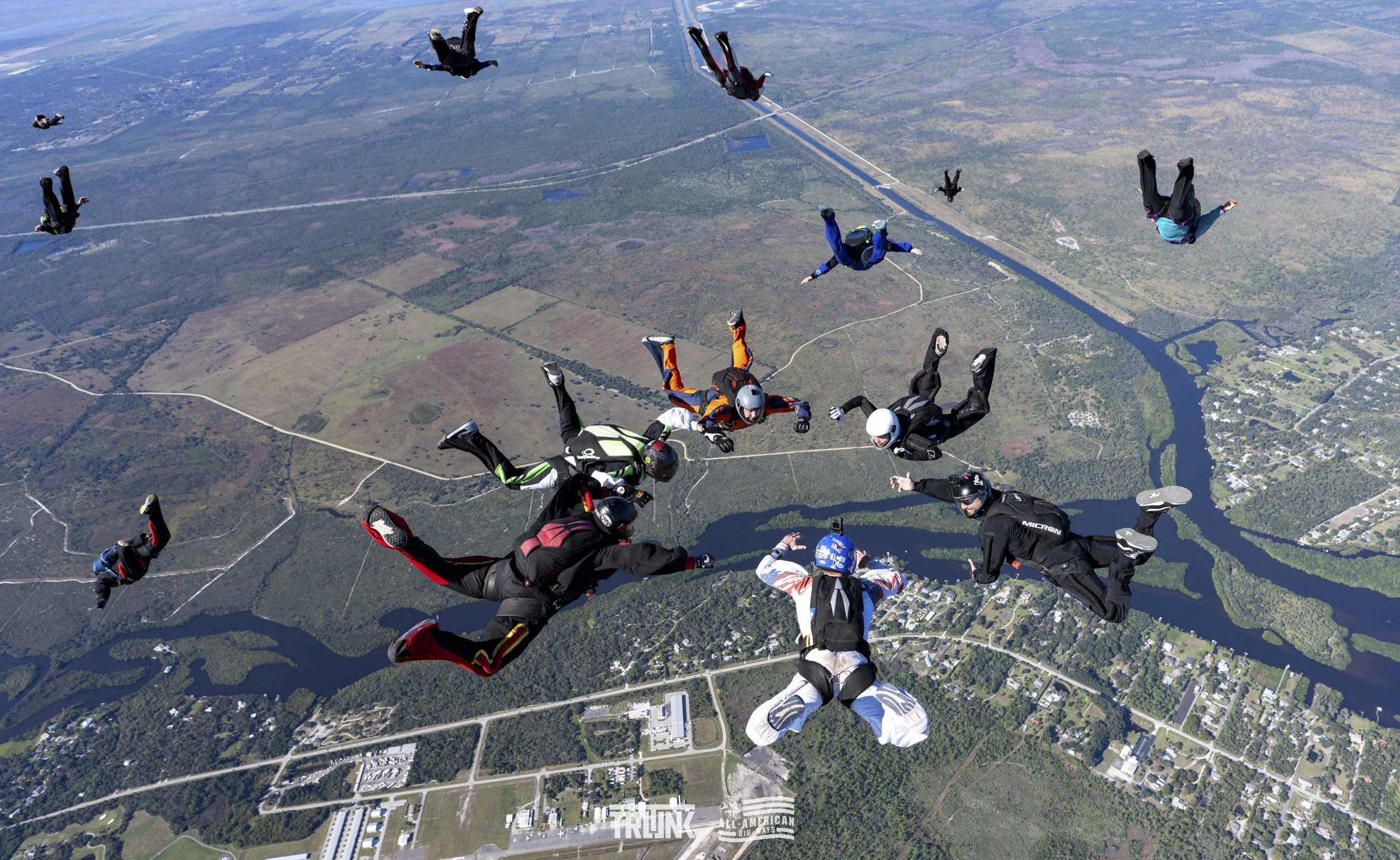 Group of tandem skydivers free-falling over a landscape with lakes, rivers, and fields.