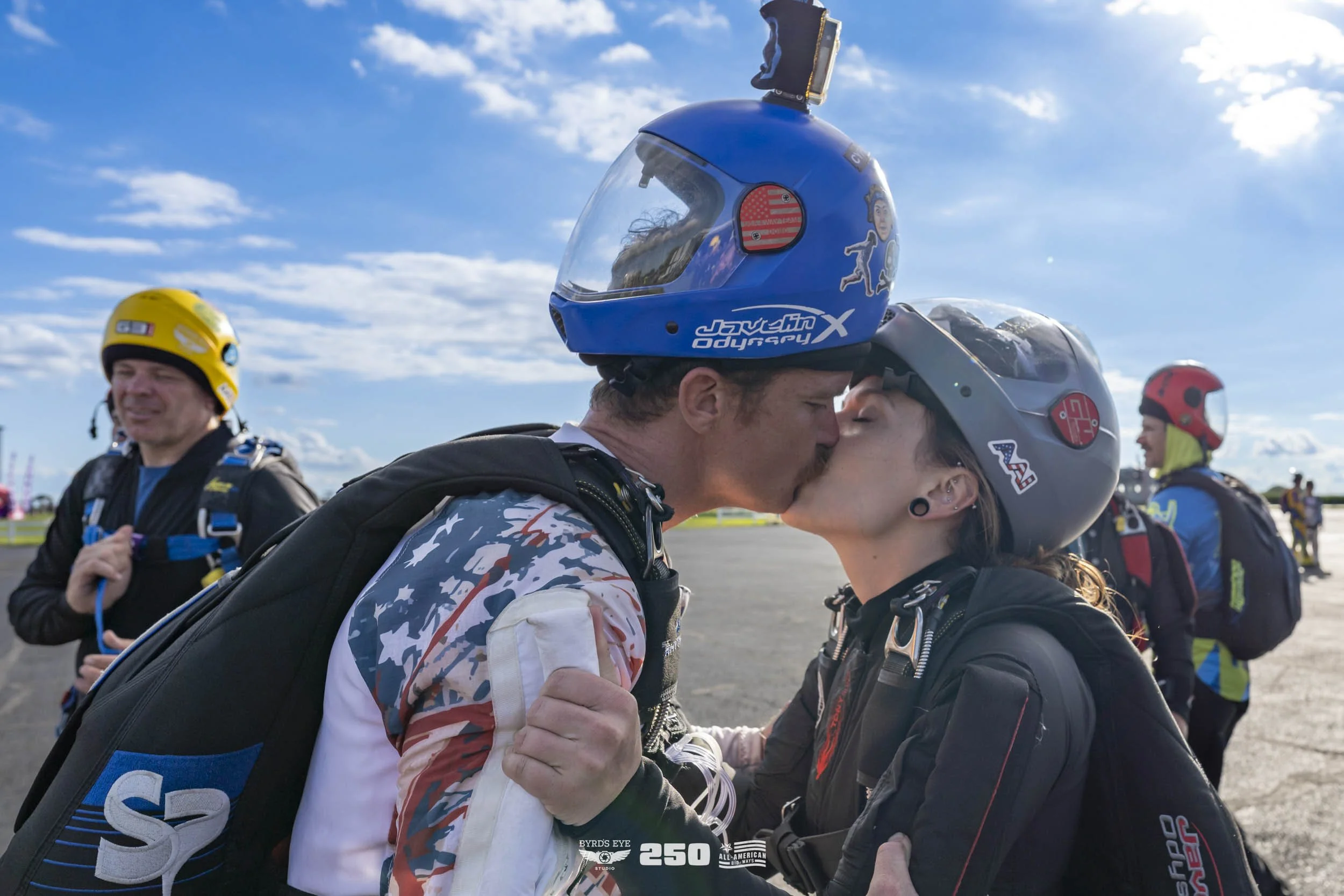 A couple kissing, wearing helmets and racing suits, at a racing event with other racers and a blue sky in the background.