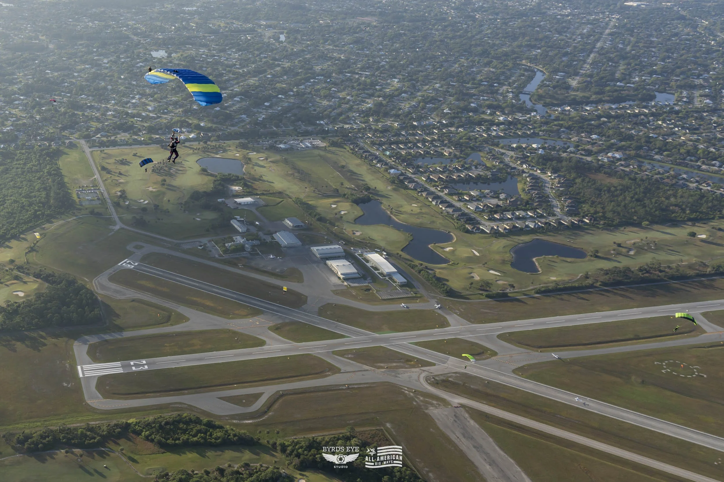 Three skydivers with parachutes descending over an airport and suburban area with lakes and houses.