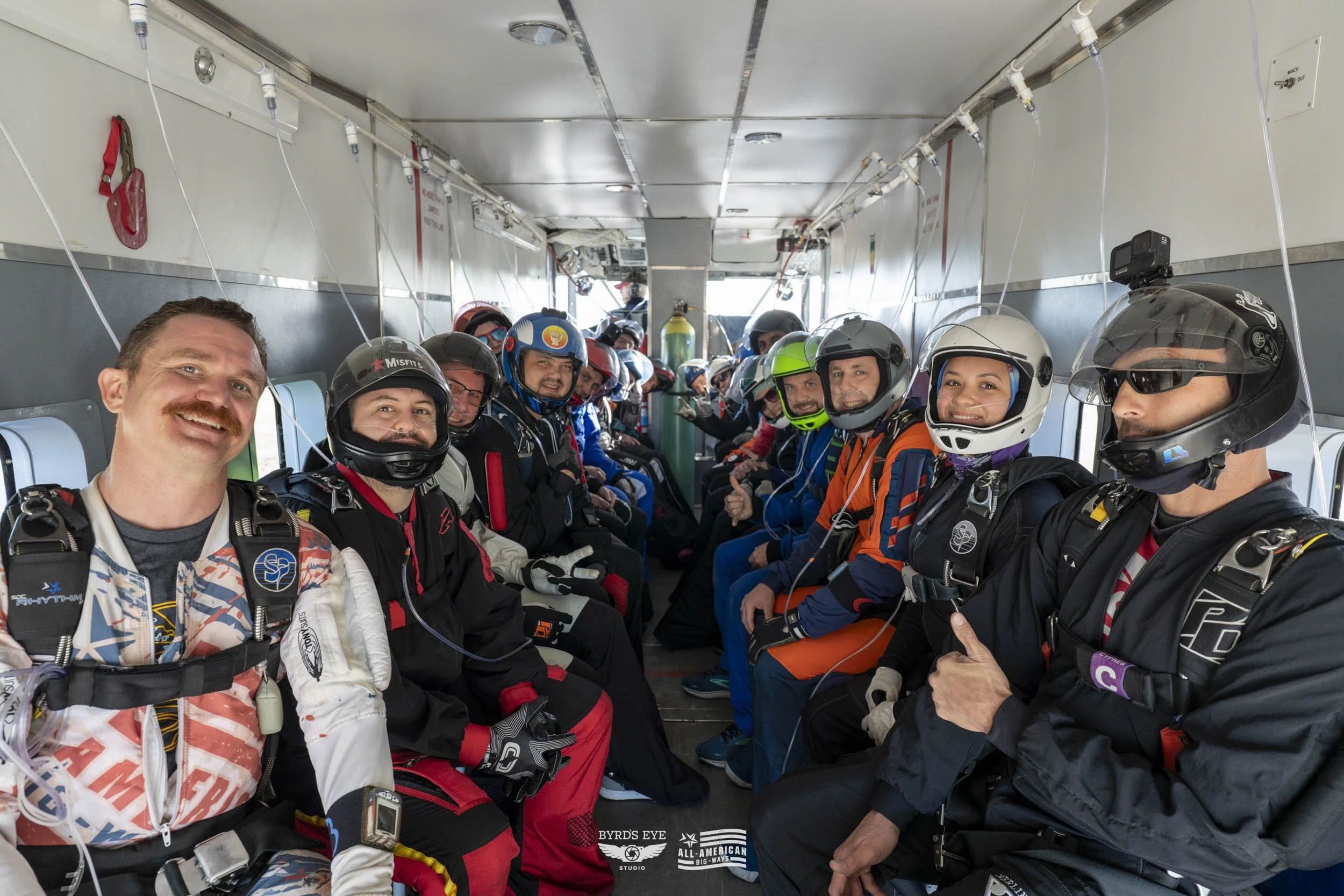 Skydivers seated inside an aircraft, wearing jumpsuits and helmets, smiling and giving thumbs-up.