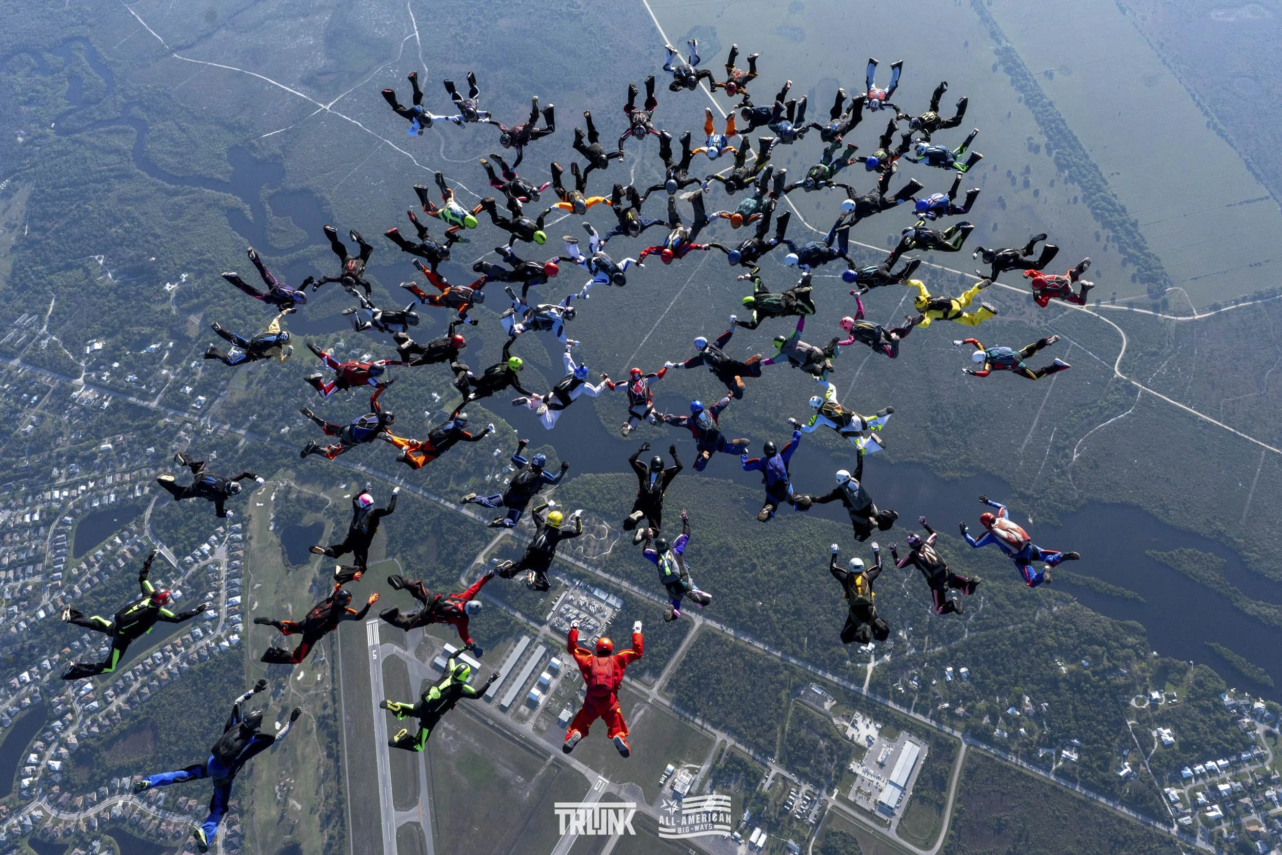 A group of skydivers in colorful jumpsuits and helmets free-falling over a landscape with lakes and roads.