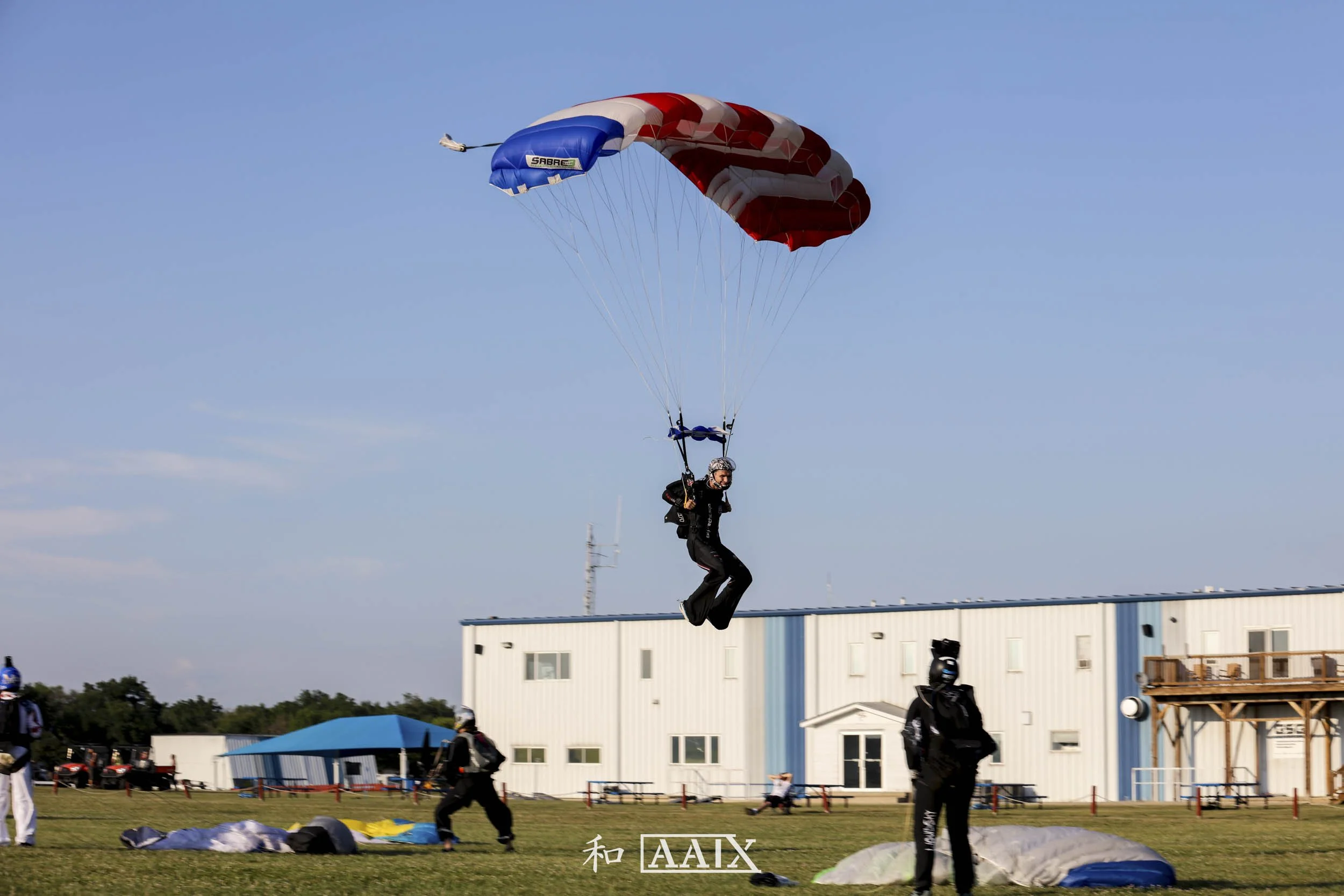 Person with a helmet and black suit parachuting down with a red, white, and blue parachute near a grassy field with several other people and equipment, a white building, and a blue sky.