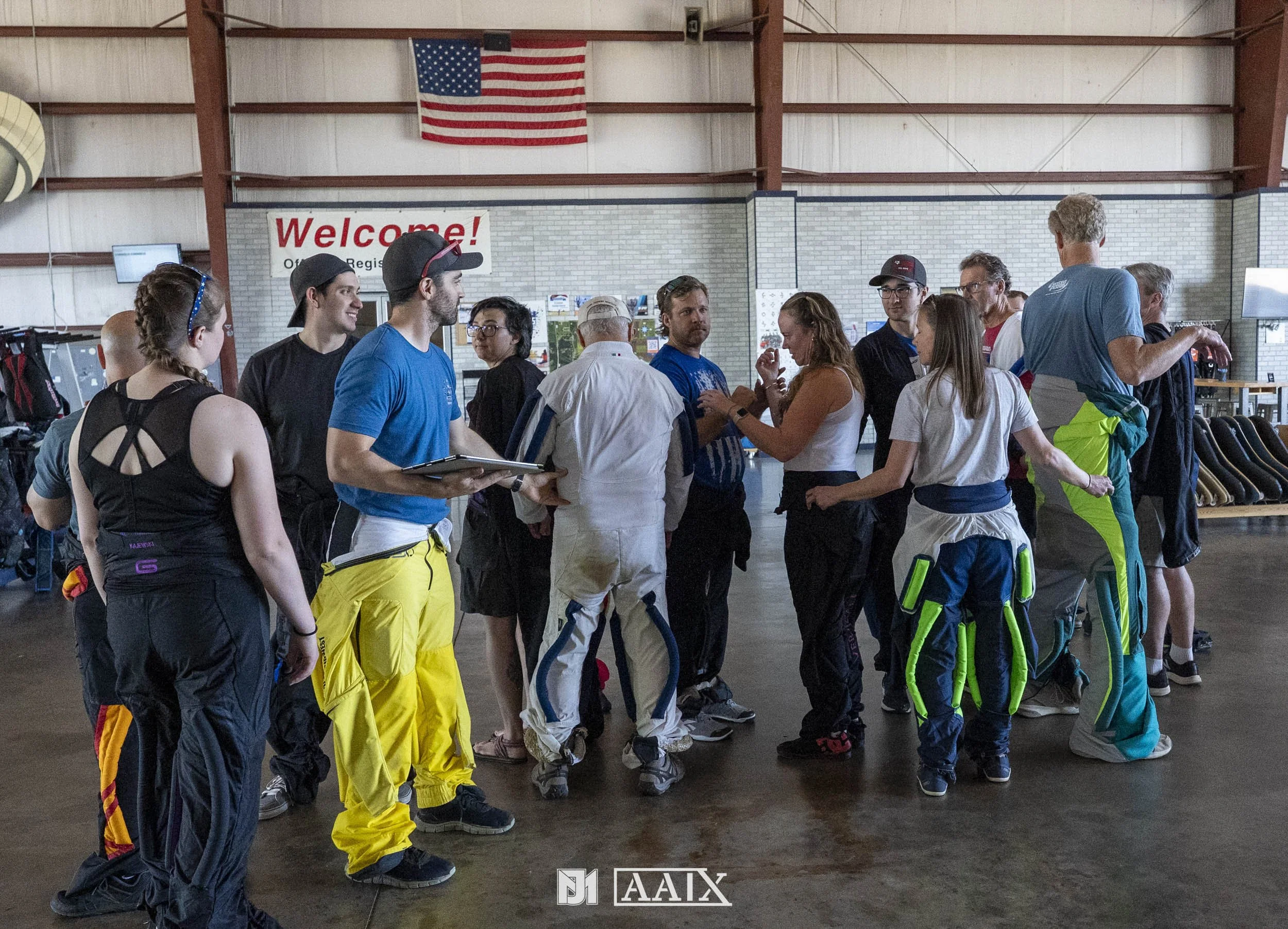 Group of people in an indoor sports facility, some dressed in athletic gear, waiting and engaging in conversation near a welcome sign.