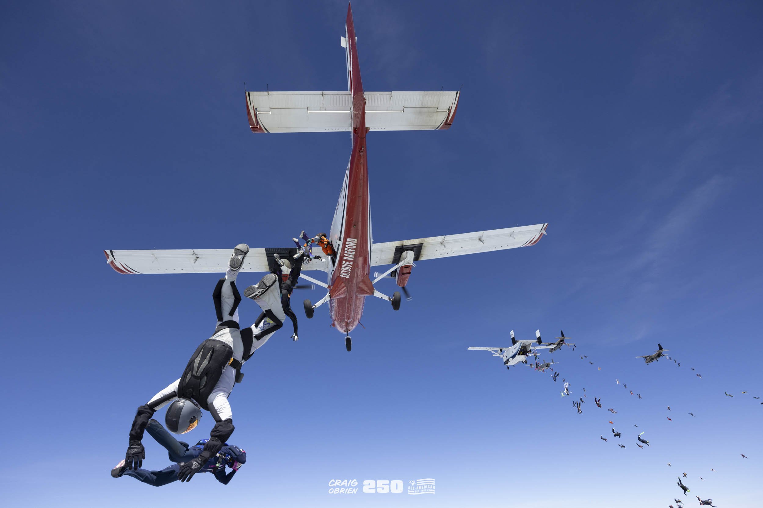 Skydivers in freefall exit an airplane against a clear blue sky, with more skydivers visible in the distance.